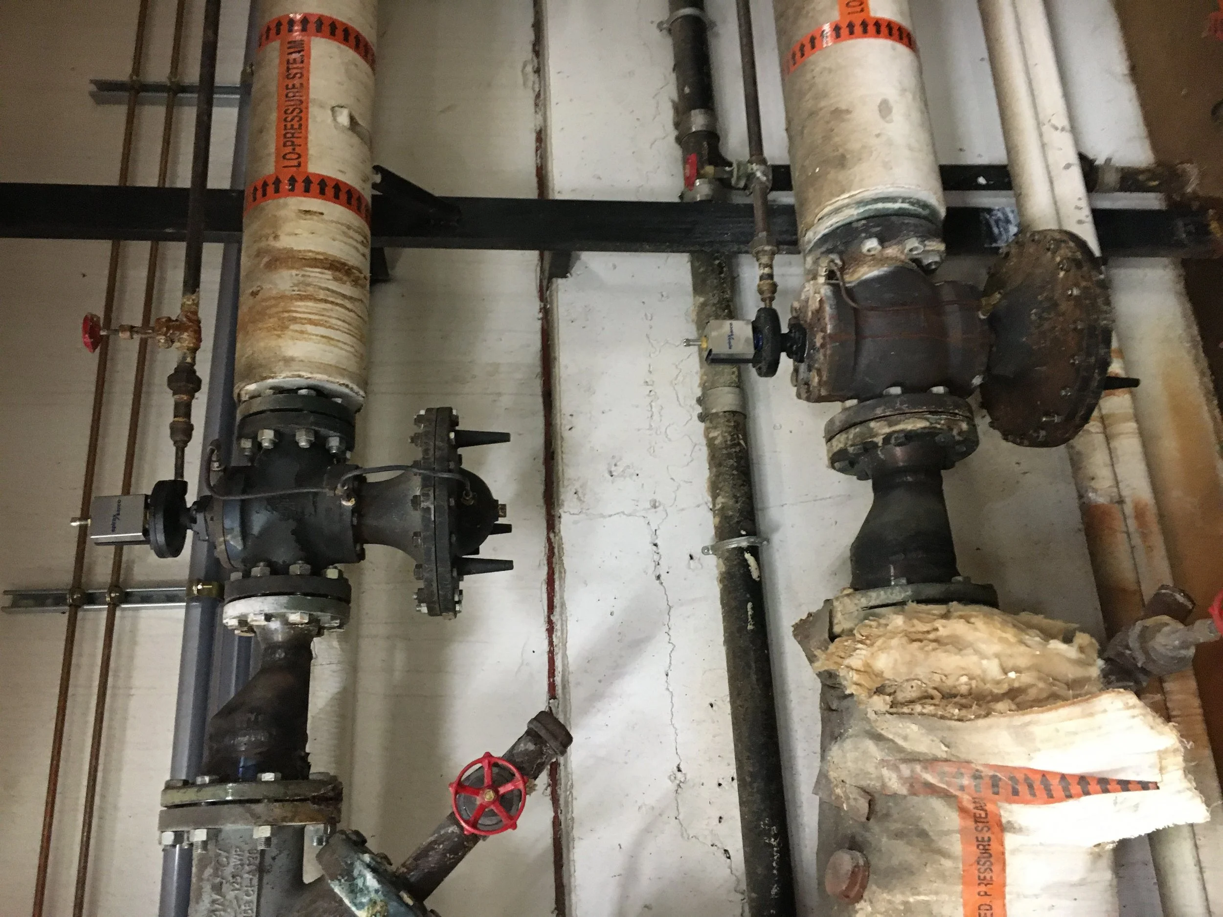 Old and rusty industrial pipes and valves with insulation, some damaged, installed on a wall with visible cracks.