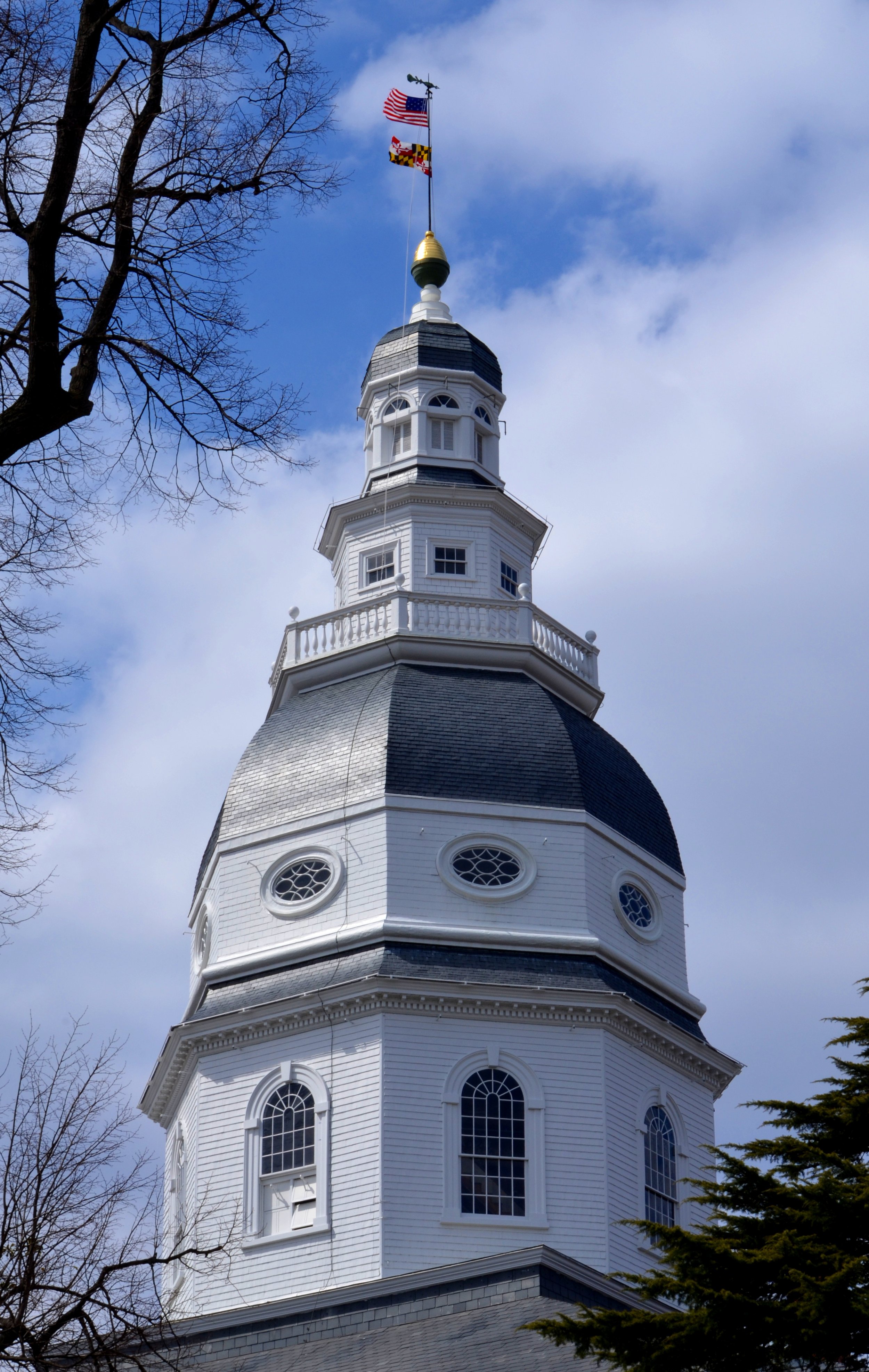 A tall white church steeple with multiple tiers, round and arched windows, and flags at the top against a cloudy sky, surrounded by leafless trees.