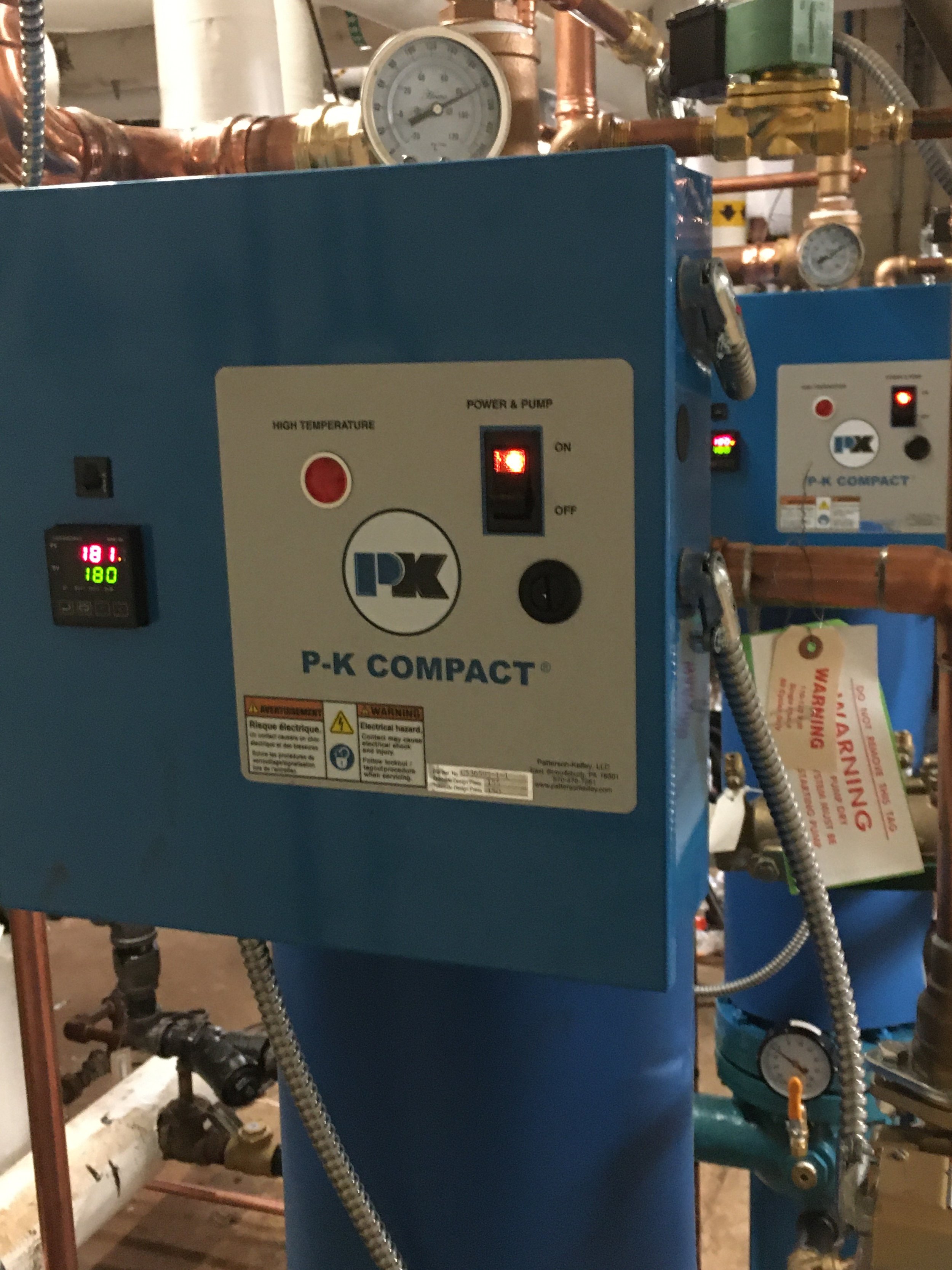 An industrial water heater labeled P-K Compact with a temperature display reading 181°F, various gauges, and copper piping.