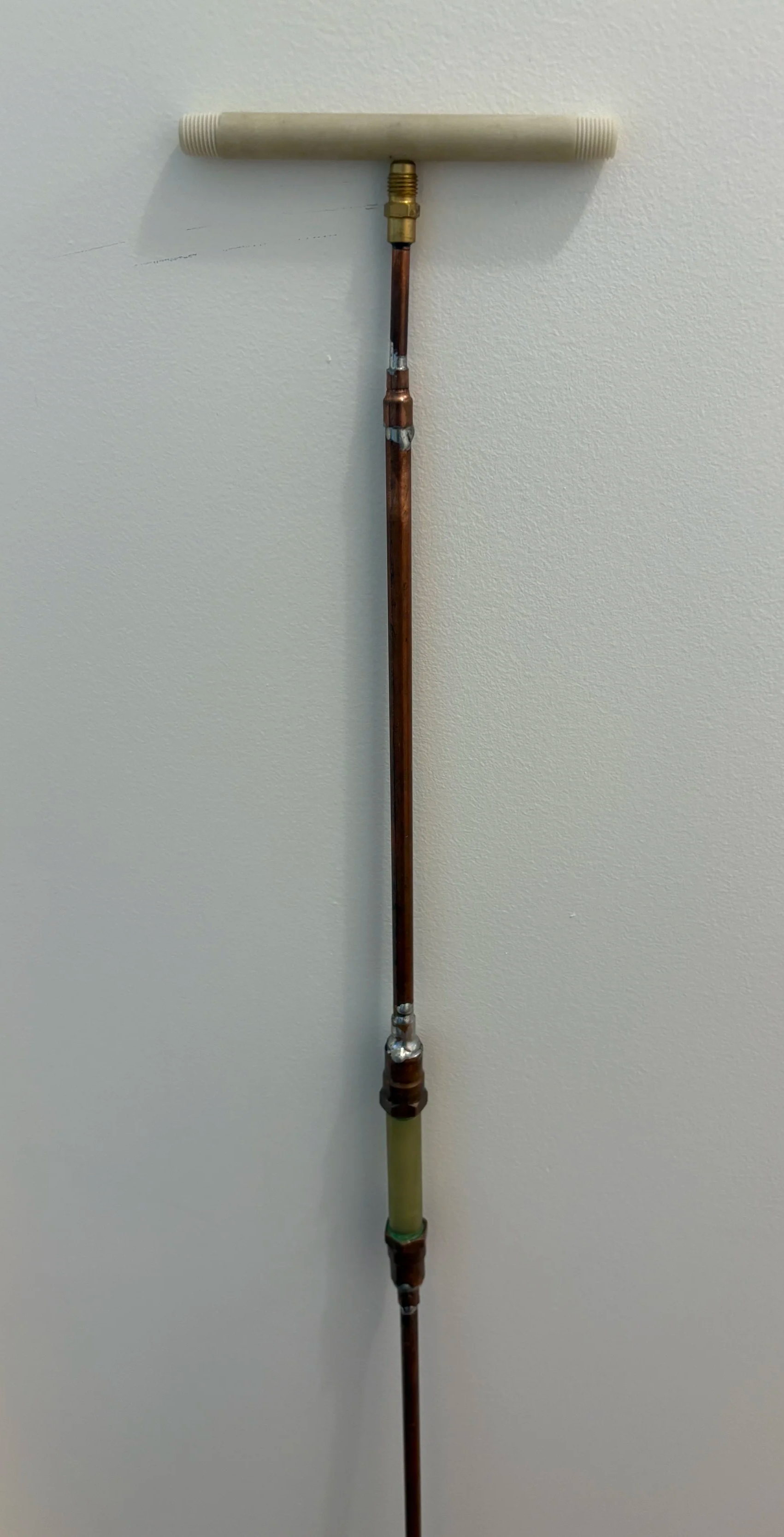 An antenna with a beige horizontal plastic rod on top, connected to a copper pipe with various fittings, mounted vertically against a plain wall.
