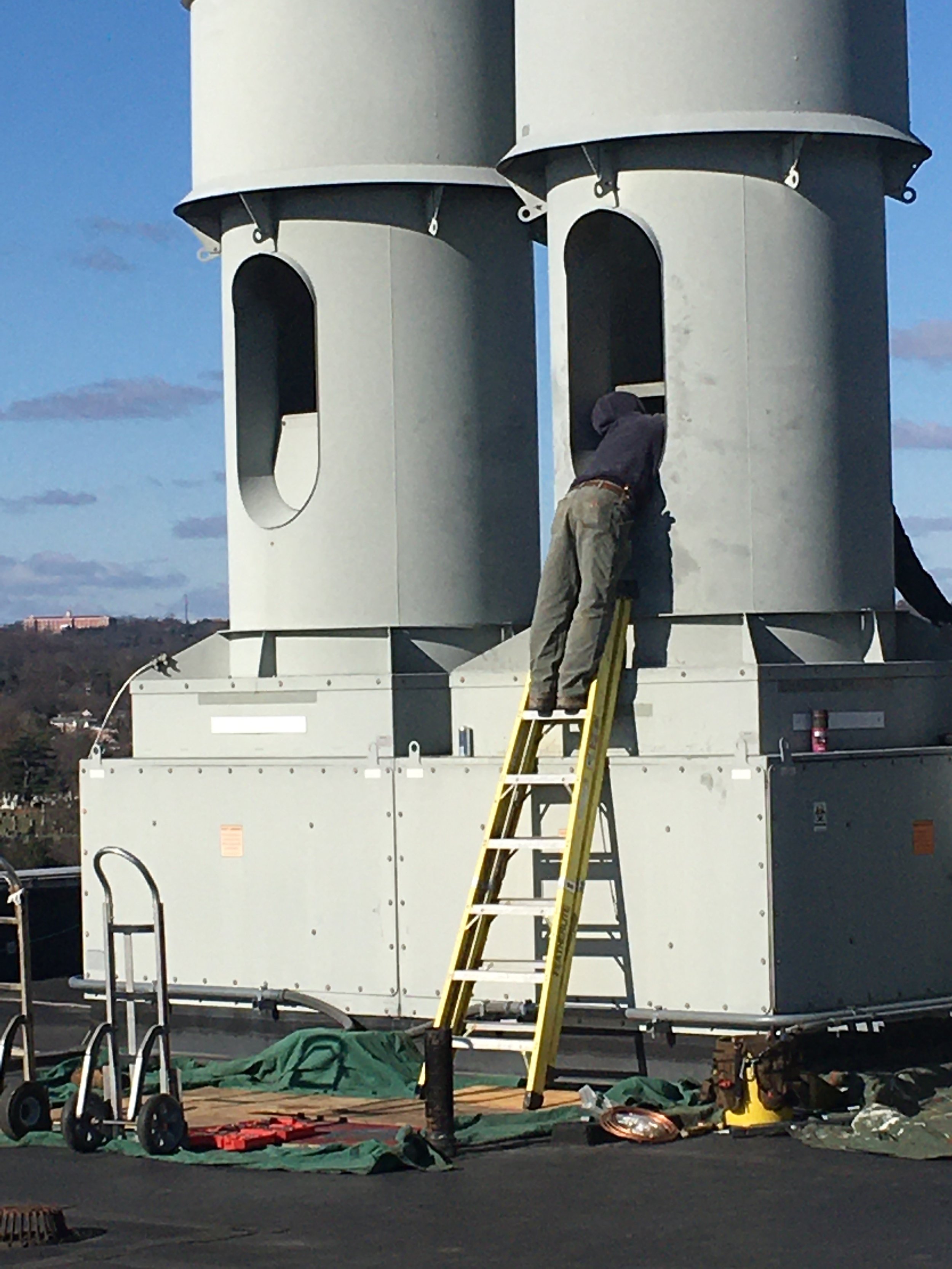 A person working on a large industrial HVAC ductwork unit, standing on a yellow ladder with tools and equipment around on the rooftop.