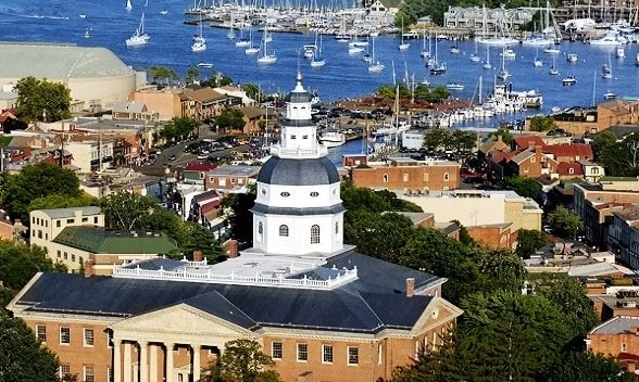 Aerial view of a lighthouse in a coastal town with boats in the harbor and colorful buildings nearby.