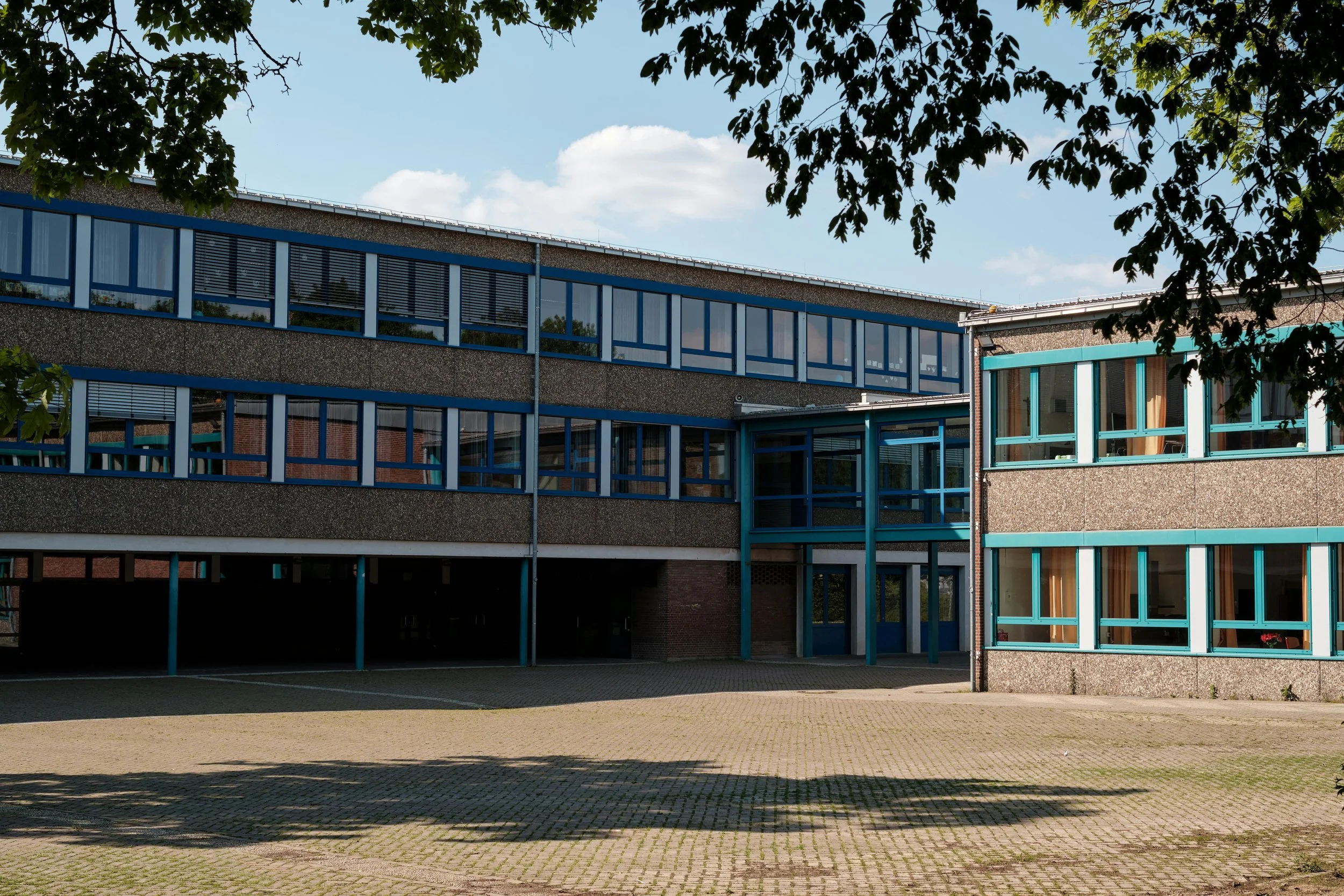 Exterior of a modern multi-story school building with large windows and a paved courtyard, partially shaded by trees.