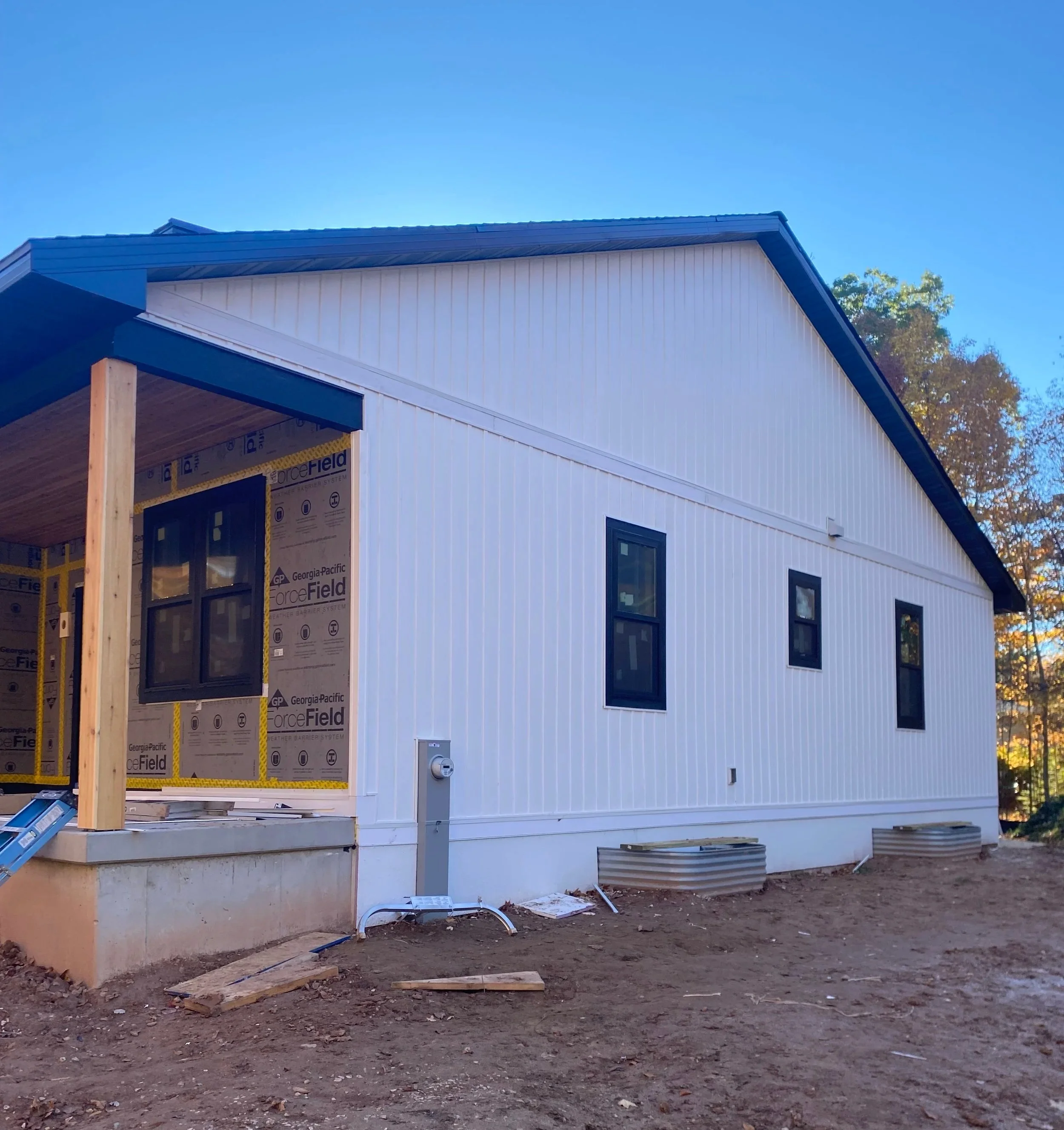 A modern farmhouse-style home under construction with white board and batten siding, black-framed windows, and a small porch area. The ground around the house is bare dirt with construction materials nearby. cedar posts. LP smart side