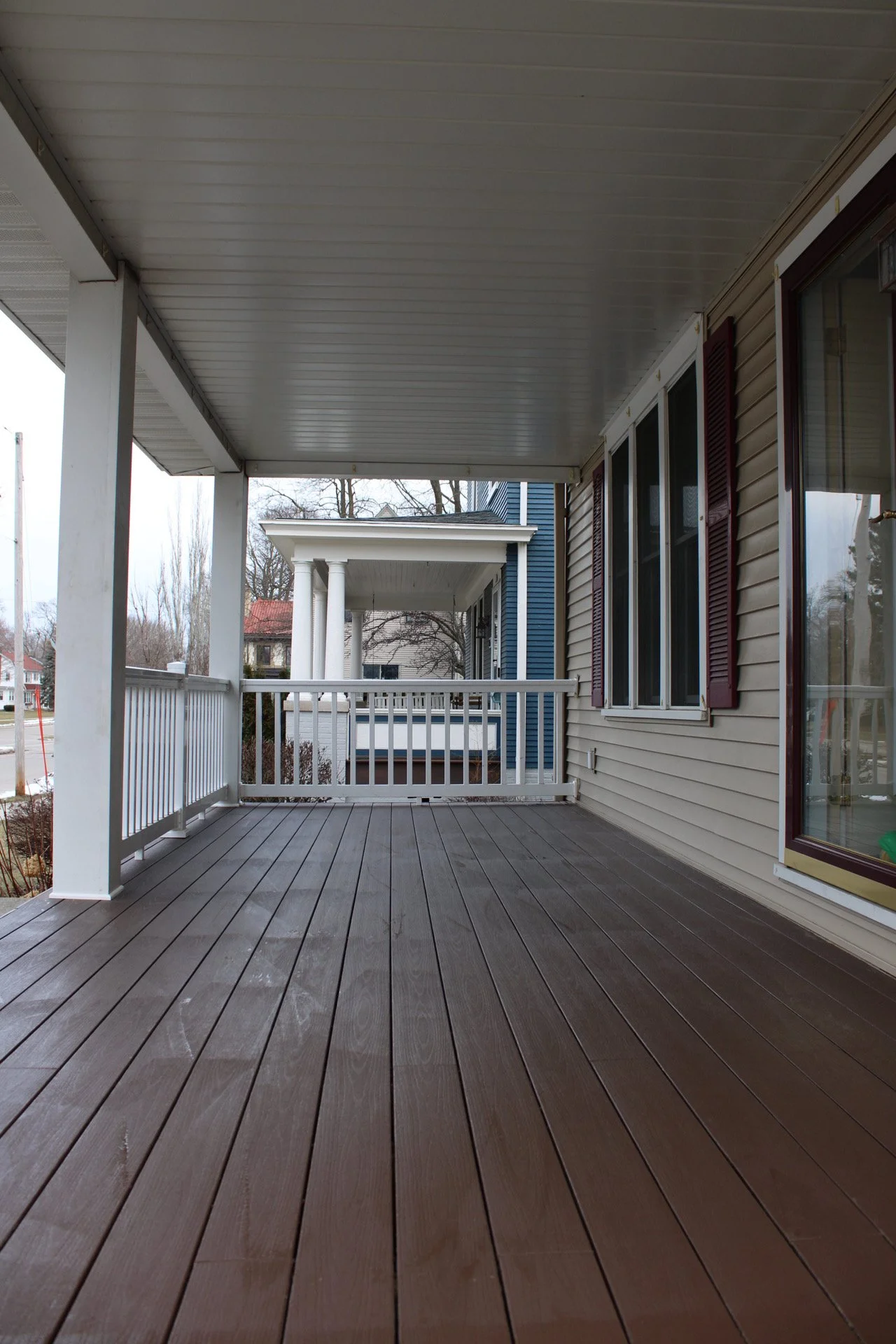 Front porch with wooden TimberTech composite decking, white railing, and part of a house with beige siding and a large window with red shutters. Updated and weather resistant 