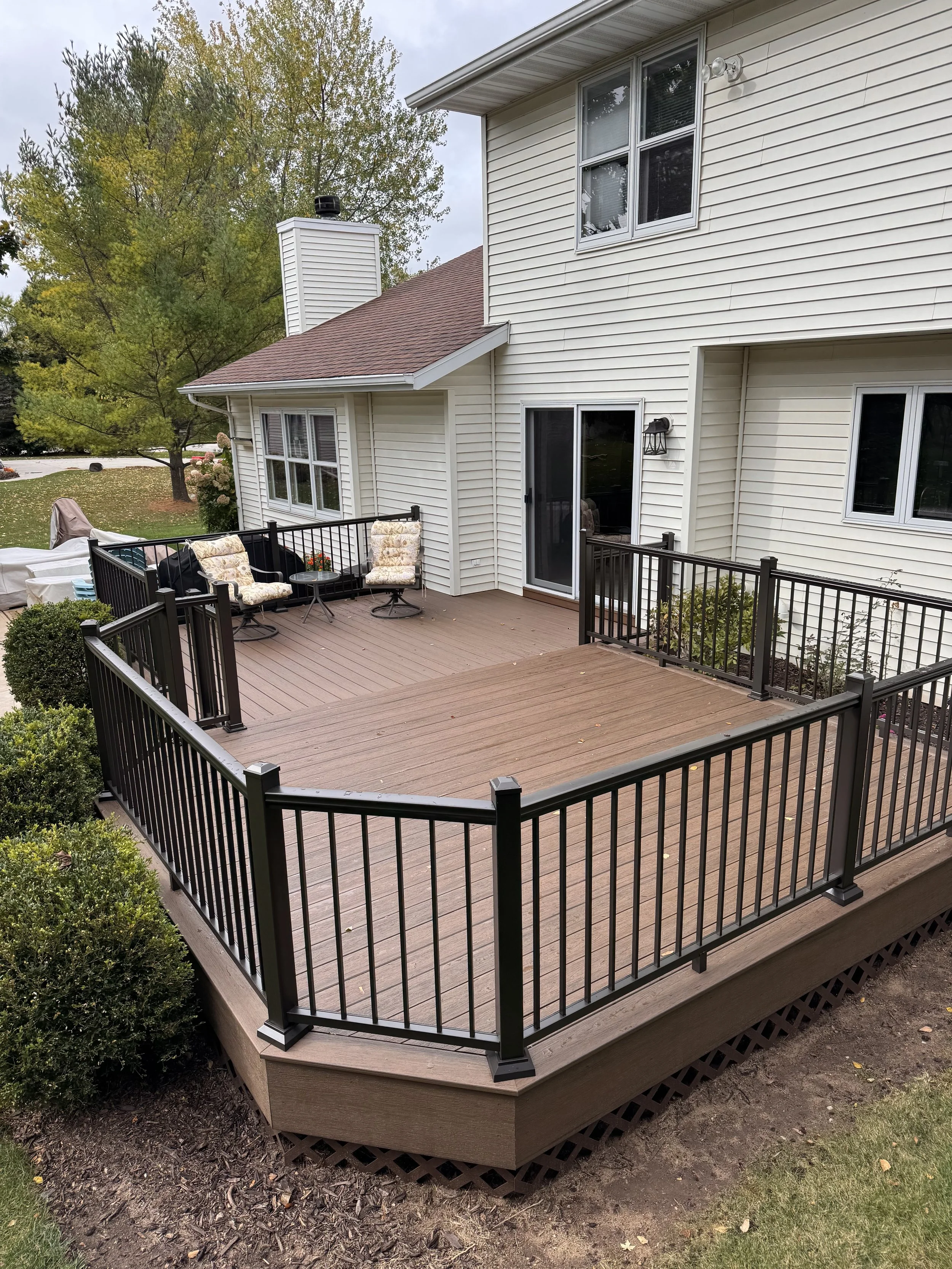 A wooden deck made of TimberTech Composite decking attached to a white house with a sliding glass door, featuring black bronze metal and aluminum railings and outdoor furniture, surrounded by trees and shrubs. custom deck