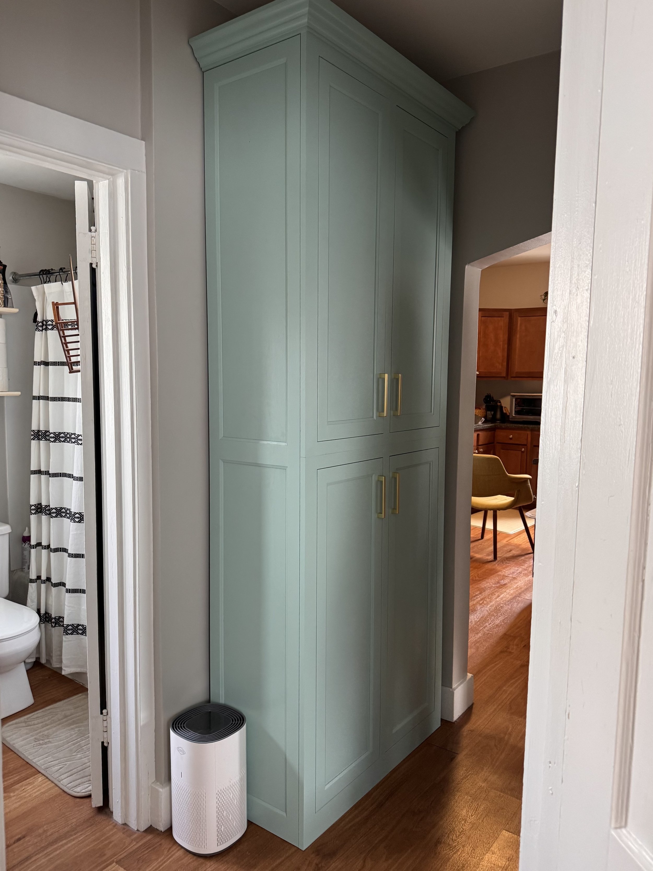 A tall, custom light blue kitchen linen closet cabinet with gold handles in a home interior, with a white air purifier at its base, adjacent to a doorway revealing a bathroom with a toilet and a shower curtain. storage, beautiful quality work