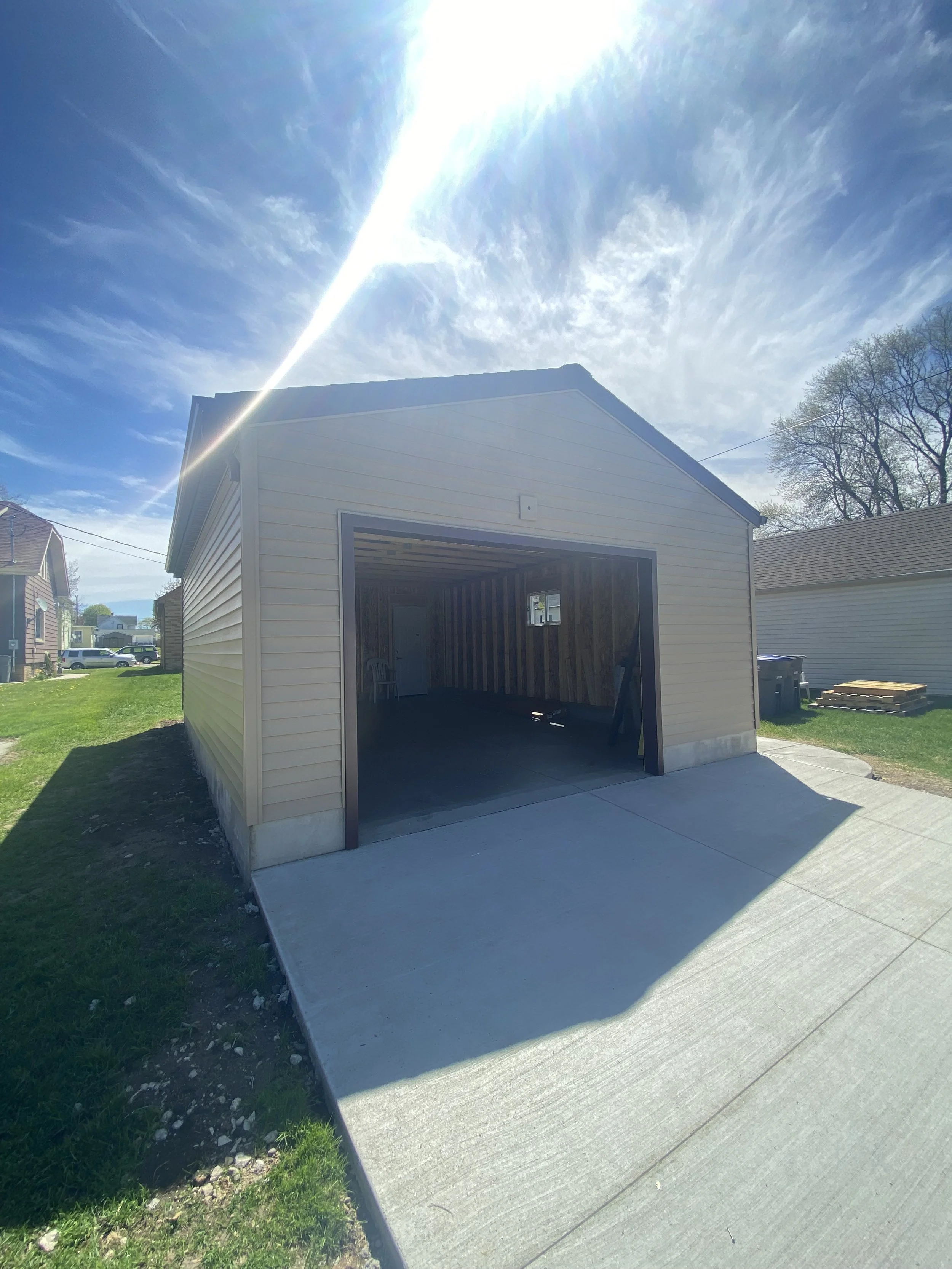 Newly constructed garage with open door, concrete driveway, grassy yard, neighboring houses, blue sky, and sunlight. fully new and updated garage for hobbies and cars