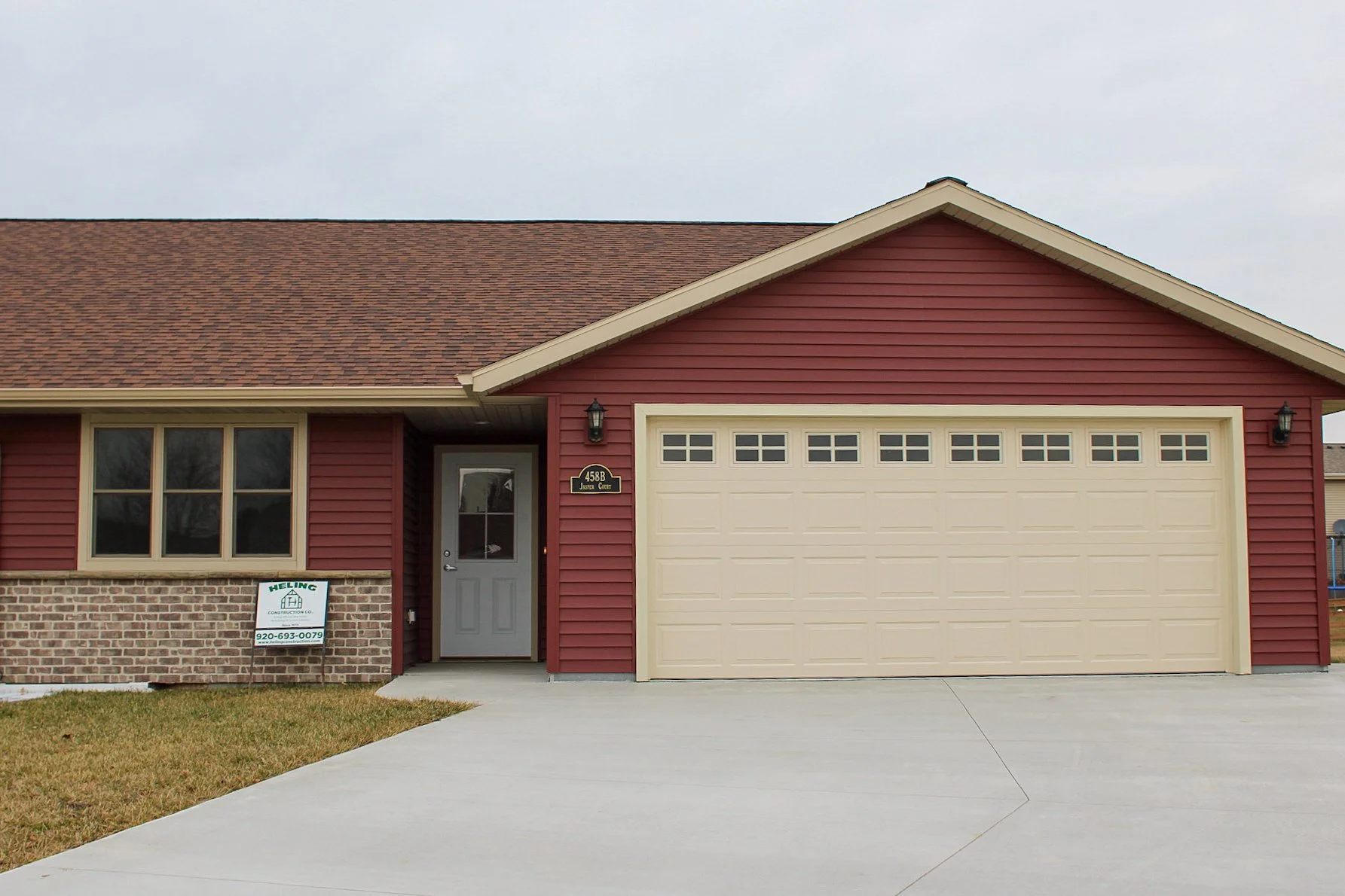 Front view of a house with a red exterior, beige garage door, and a small porch area. a custom built condo home, energy efficient. fresh concrete driveway. vinyl siding