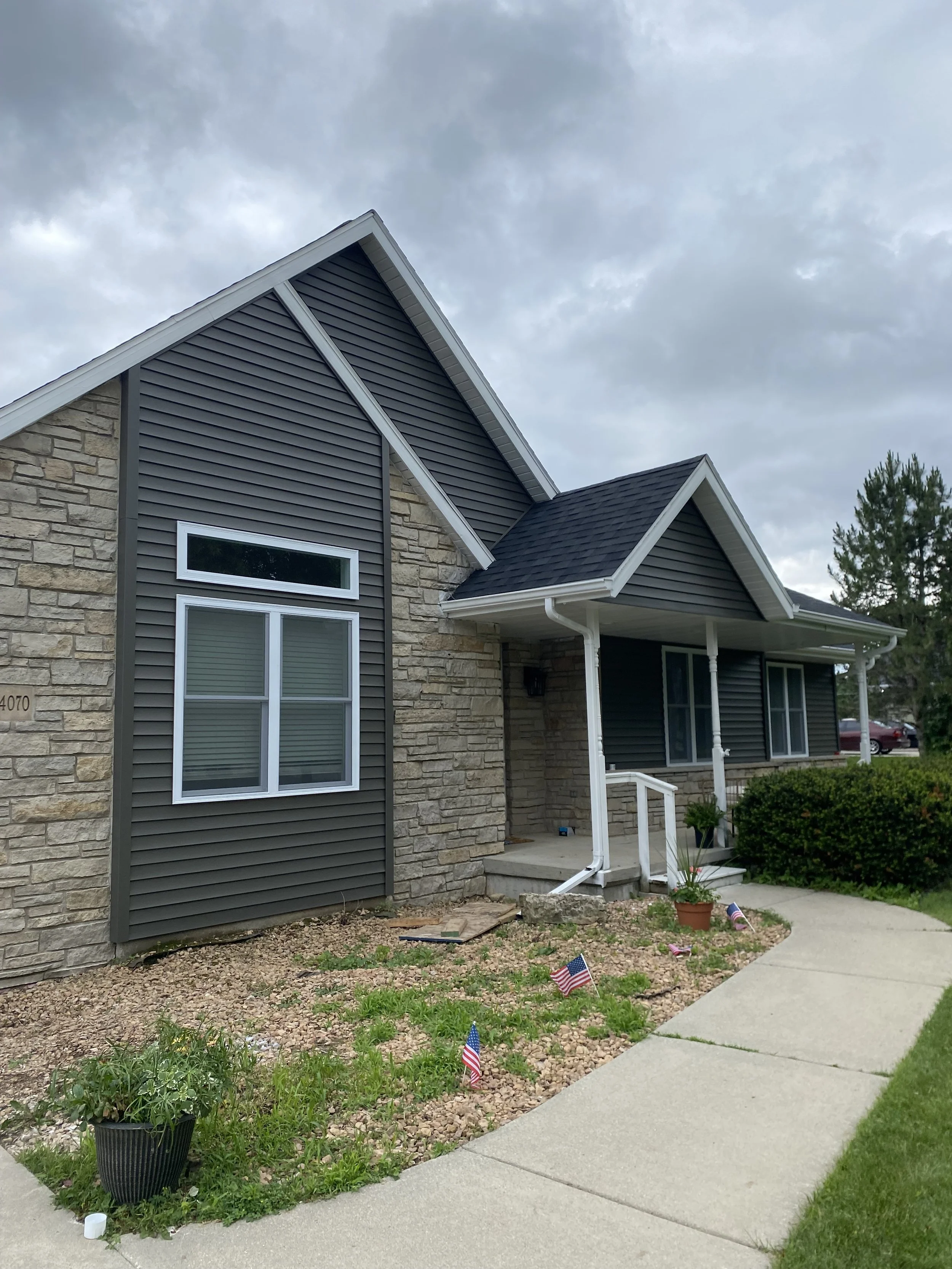 Front view of a modern house with stone and dark gray vinyl siding, a small front porch, and a landscaped yard with grass, small plants, and American flags. Concrete walkway.