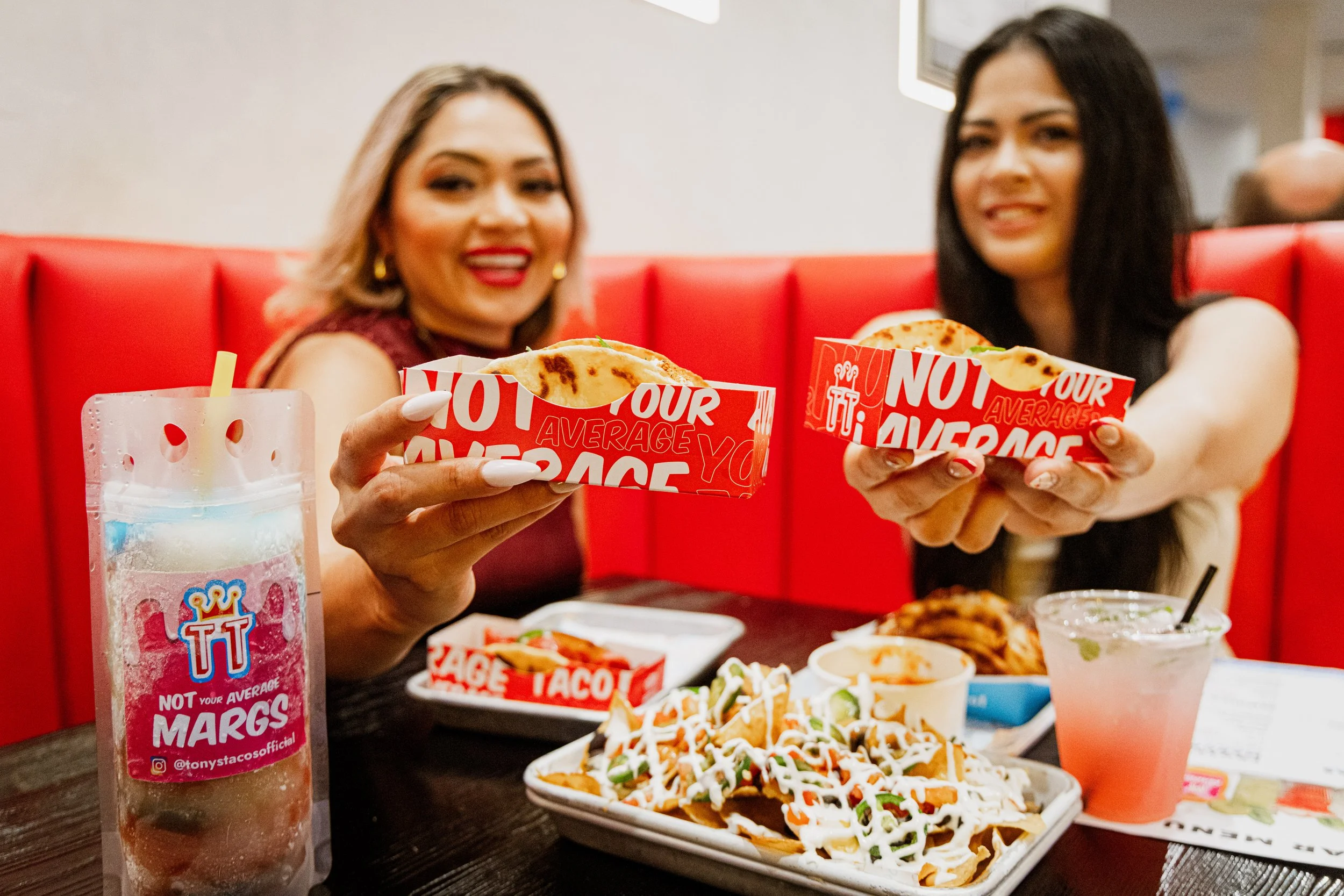 Two women sitting at a table in a restaurant, holding tacos in red and white containers, with nachos and drinks on the table.