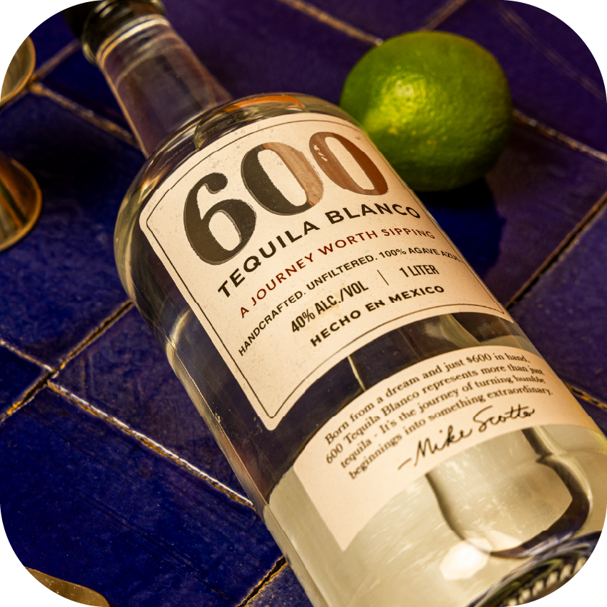 A bottle of 600 tequila blanco lying on a blue tiled surface with a lime nearby.