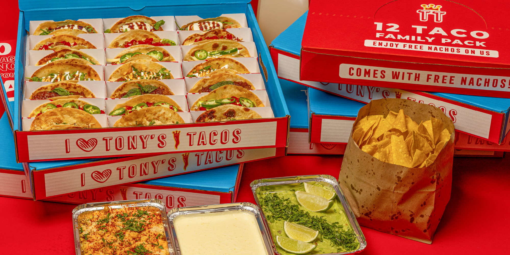 Multiple boxes of Tony's Tacos Taco Boxes with various taco fillings, a paper bag of tortilla chips, and containers of guacamole, cheese dip, and salsa on a red table.