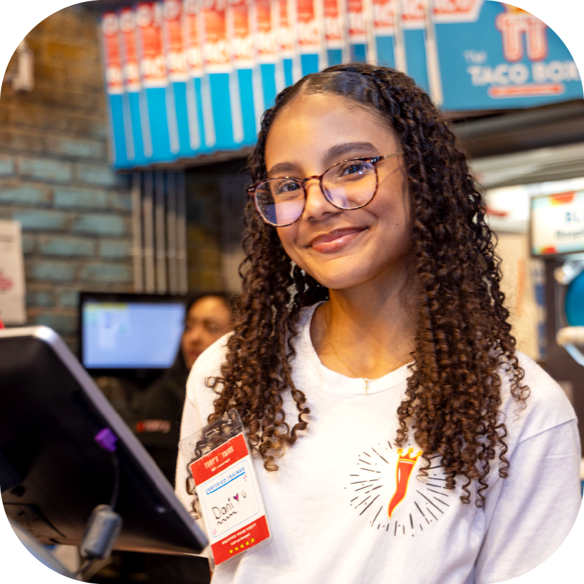 Young woman with curly hair and glasses smiling at the checkout counter of Tony's Tacos, wearing a white shirt with a Tony's Tacos logo, in a fast food restaurant.