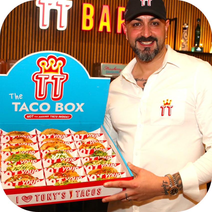 A man smiling and holding a Tony's Tacos Taco Box. The background has wooden panels and bright signs, including the words 'TT Bar'.