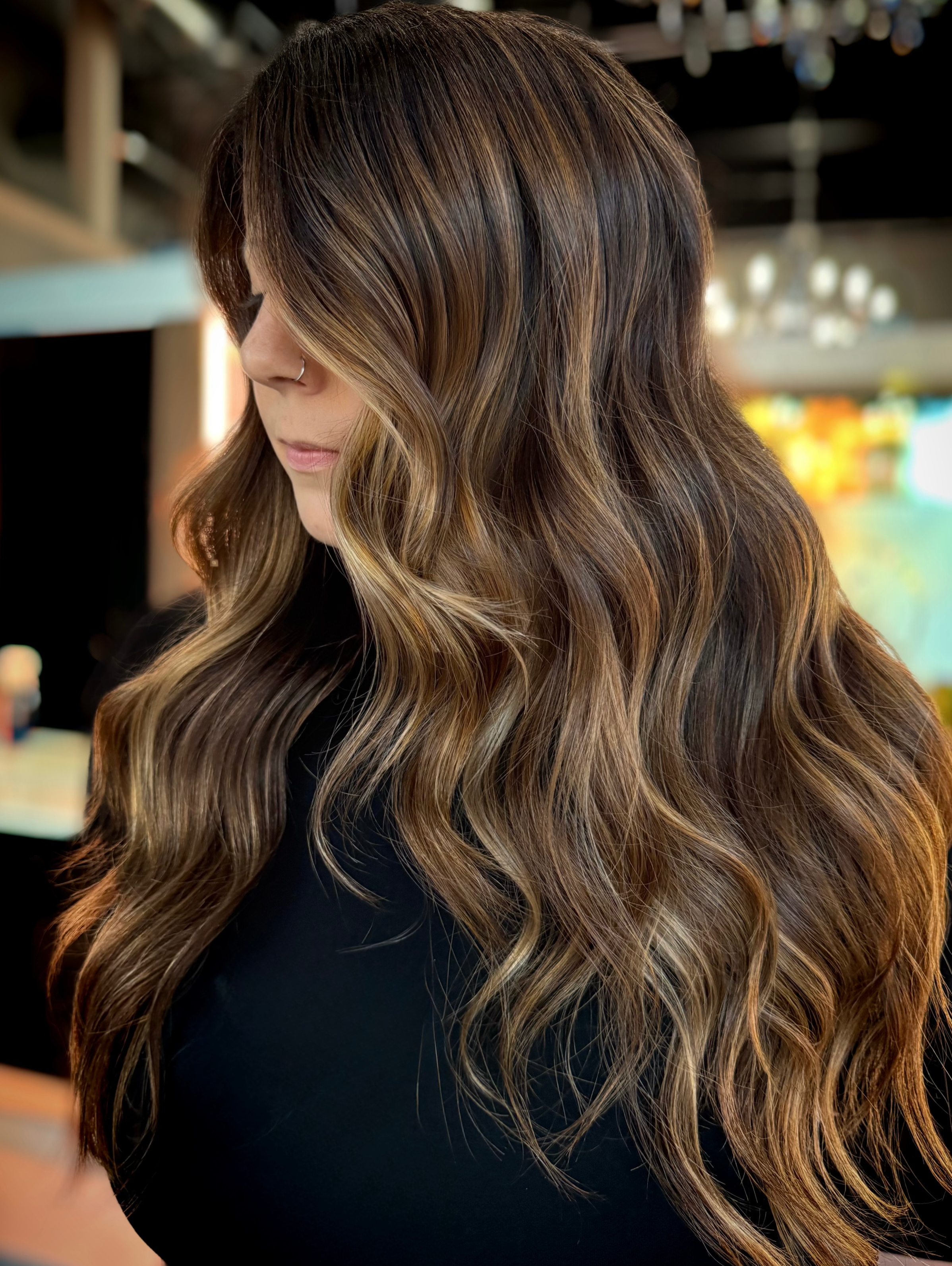 Close-up of a woman with wavy, honey-blonde hair and a nose ring, looking down, blurred background.