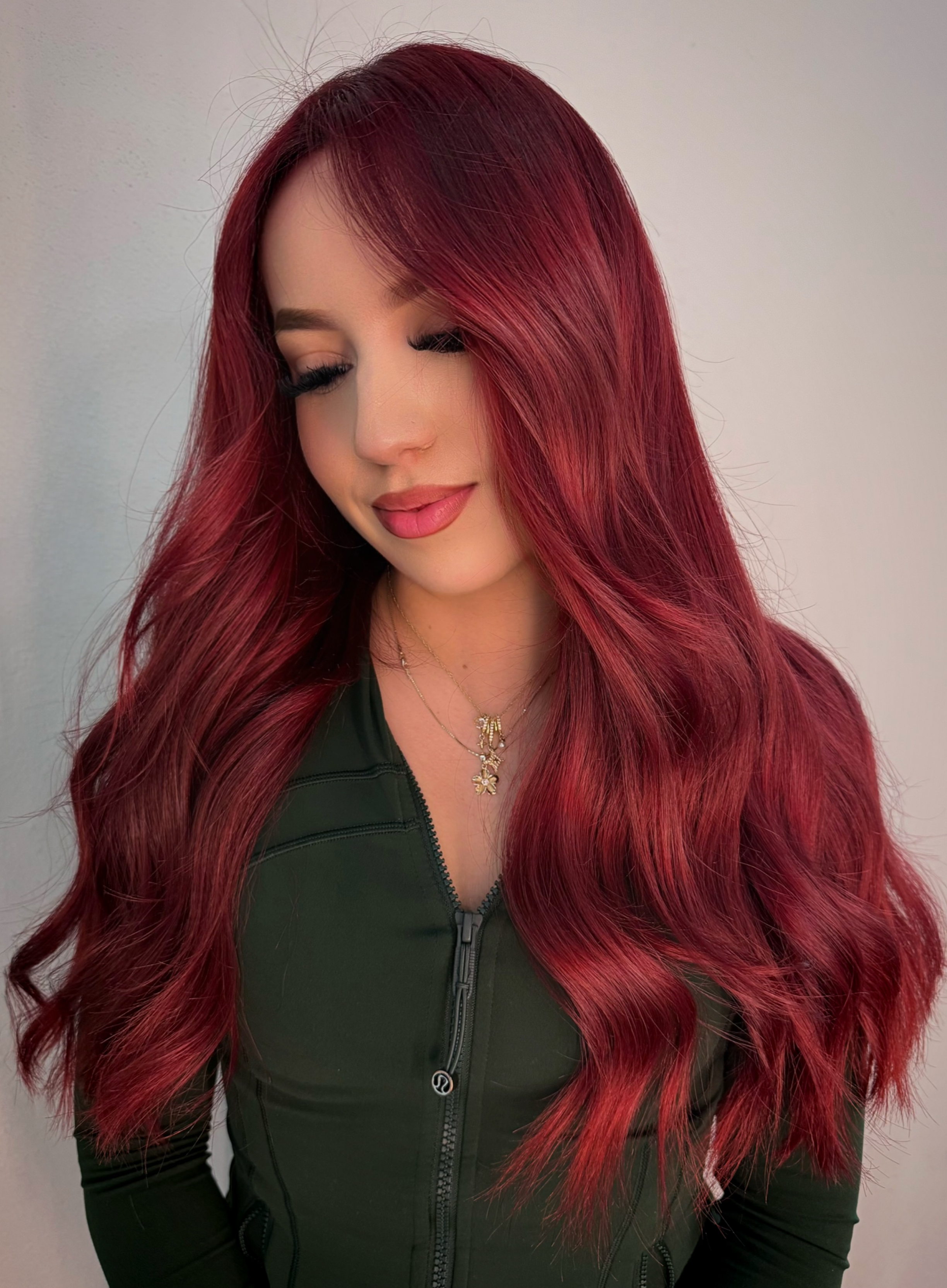A woman with long, wavy, red hair, wearing a green zip-up jacket and a gold necklace, with her eyes closed and a slight smile.