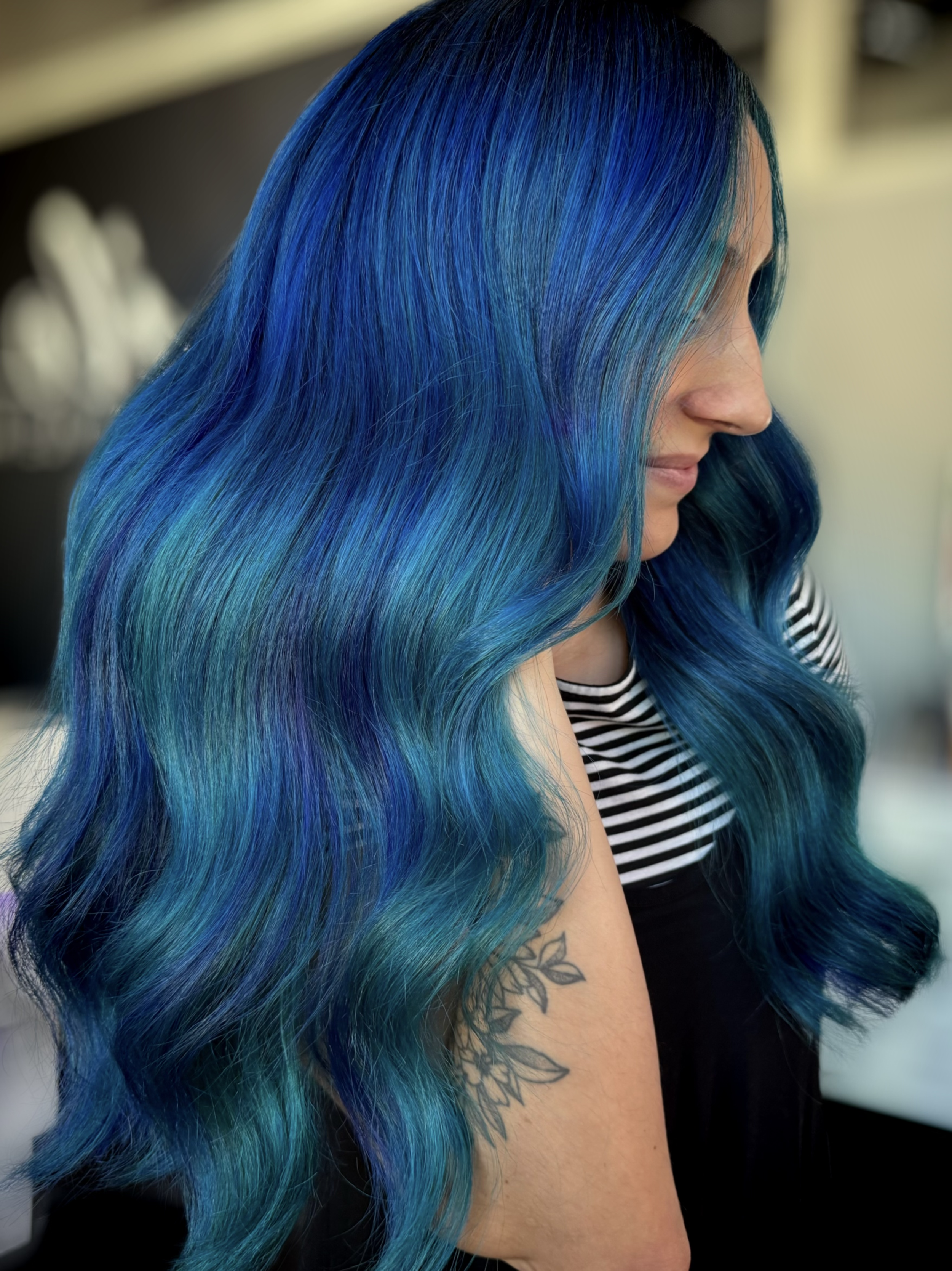 A woman with long, wavy hair dyed in shades of blue and teal, wearing a black and white striped shirt, and a tattoo of flowers on her arm.