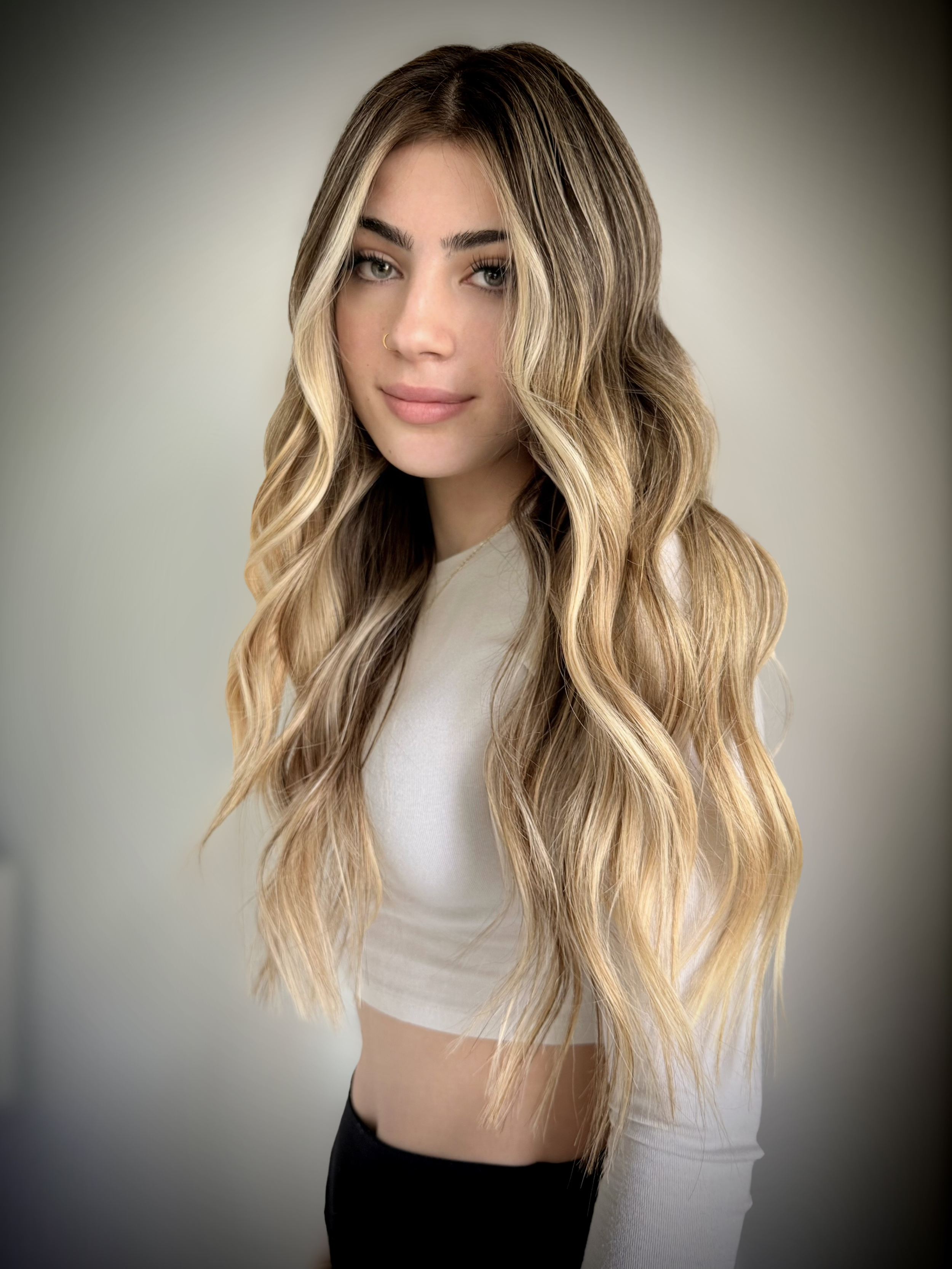 Young woman with long, wavy blonde hair, wearing a white top with long sleeves, looking at the camera with a neutral expression against a plain background.
