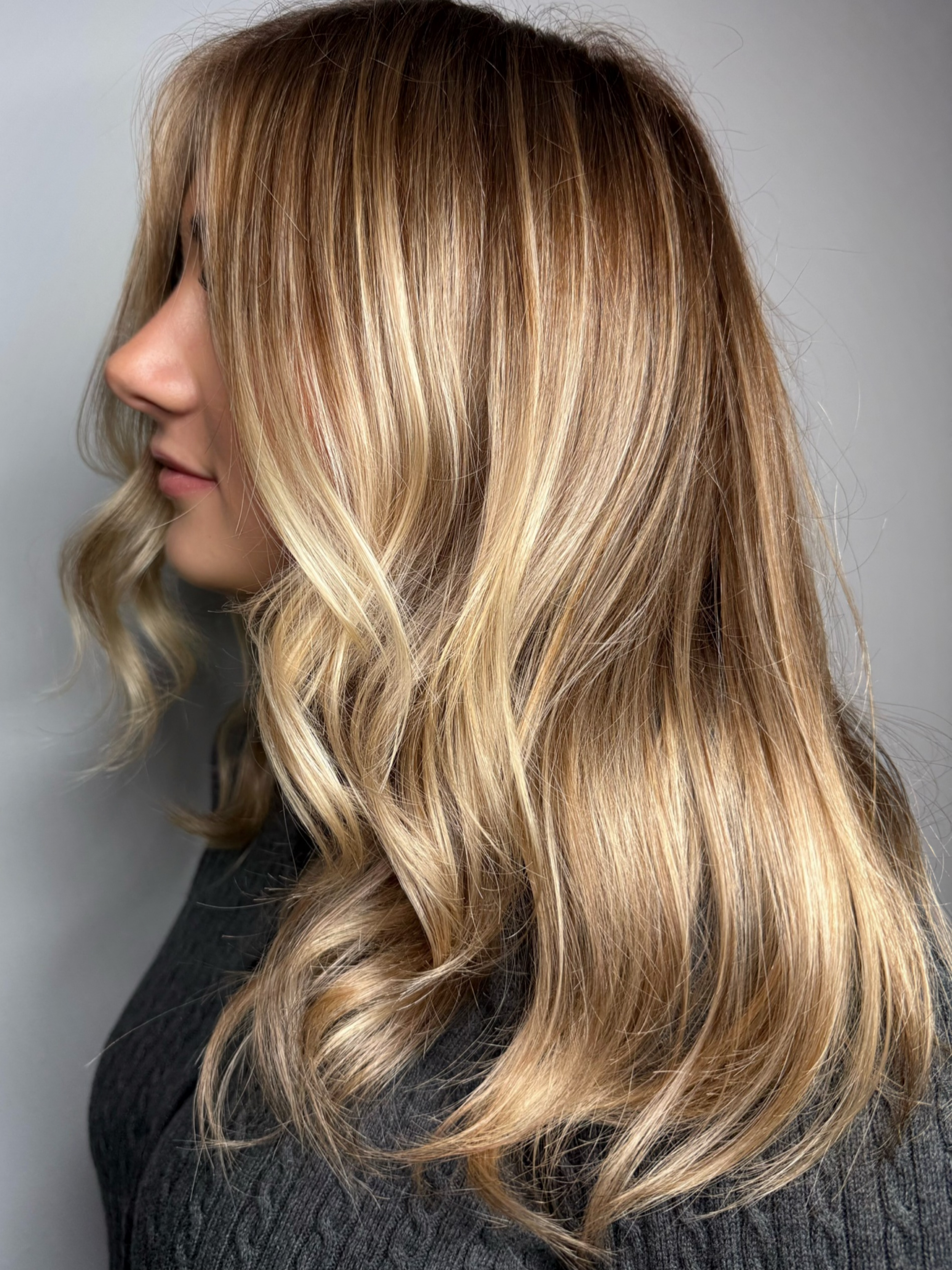 Woman with long, wavy, blonde hair with darker roots, wearing a dark sweater.