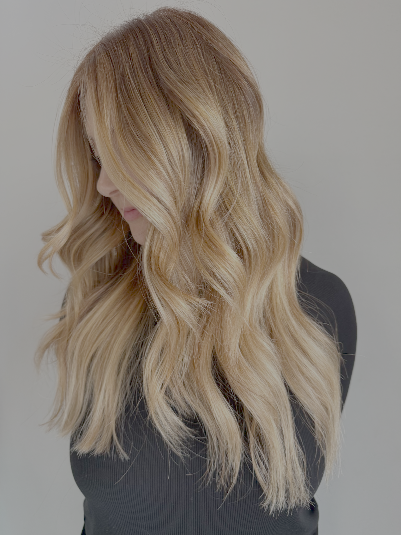 A woman with long, wavy blonde hair wearing a black top. Her head is tilted downward, and her hair covers part of her face. The background is plain light gray.