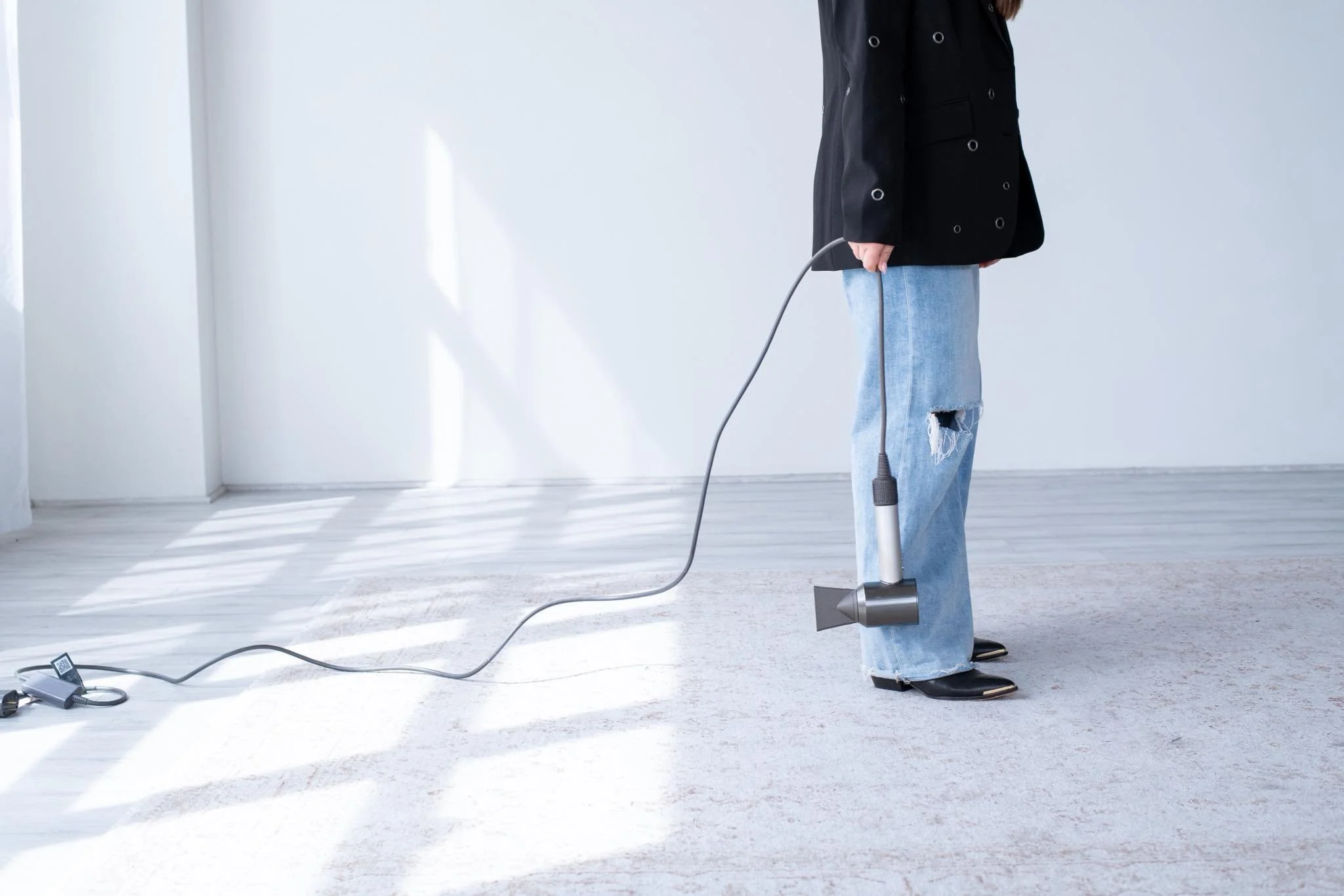 Person wearing a black coat and ripped jeans vacuuming a white carpet in a bright, modern room.