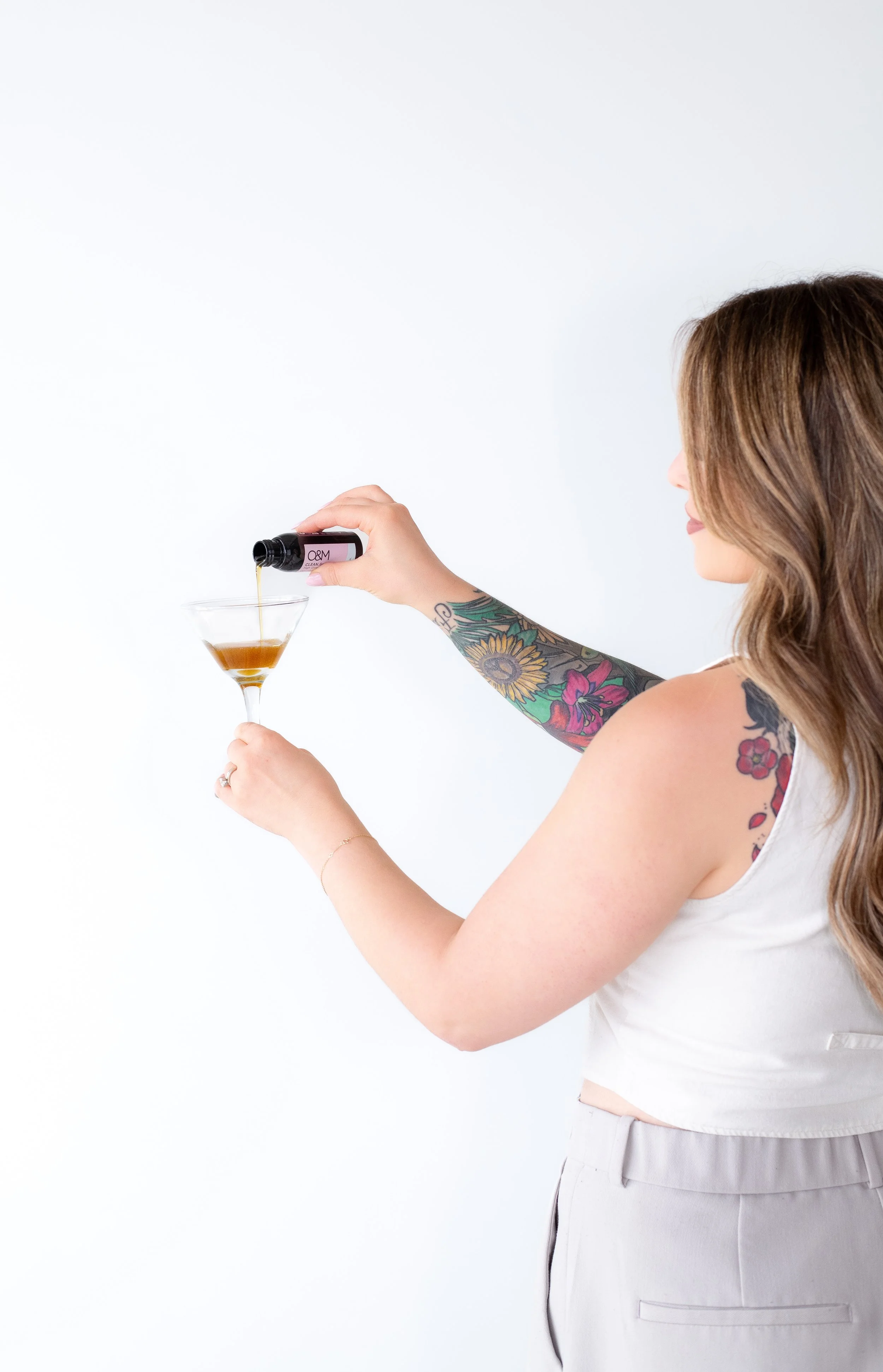 A woman with colorful tattoos on her right arm is pouring a brown liquid from a black bottle into a martini glass against a white background.
