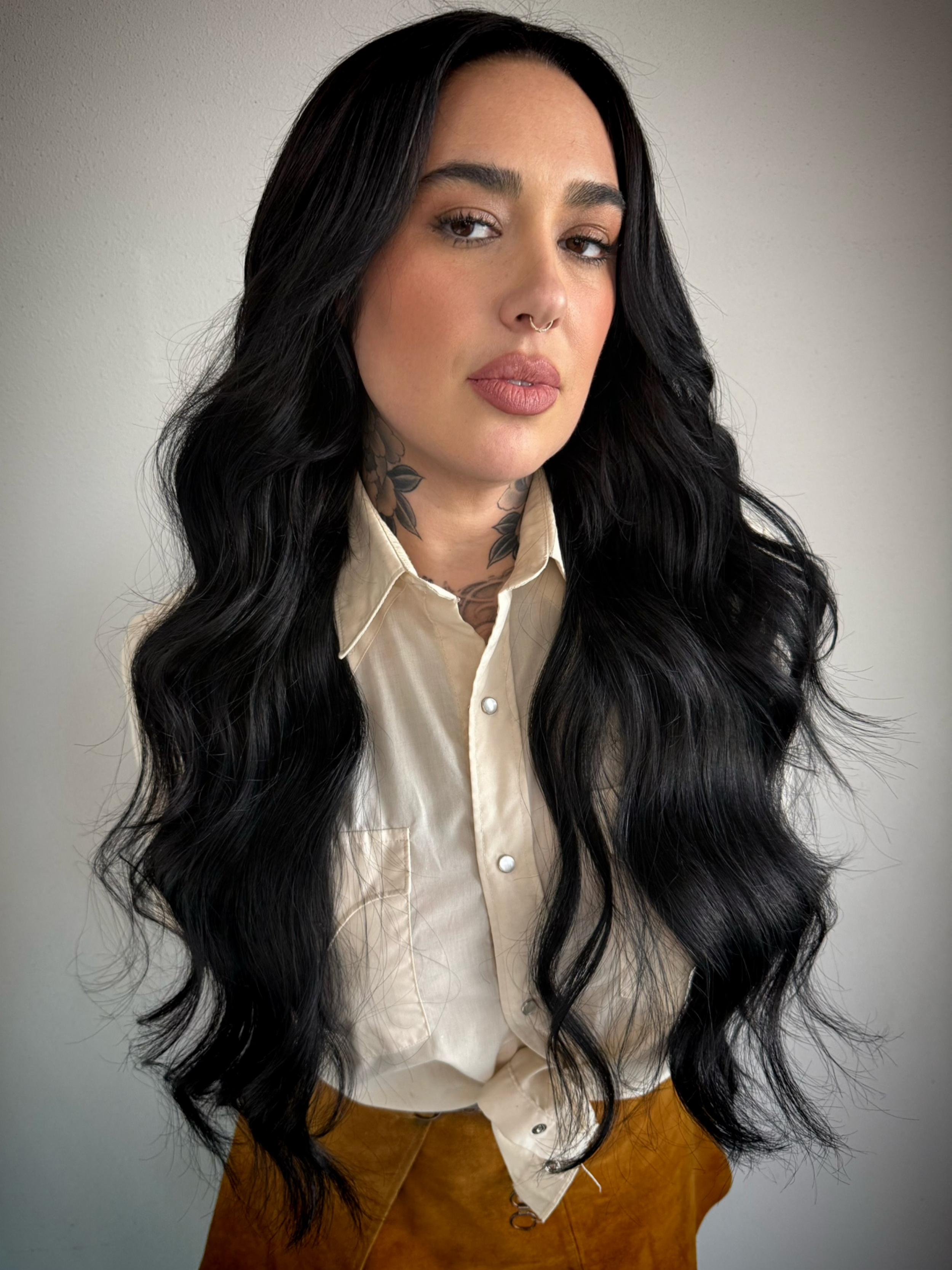 A woman with long wavy black hair, tan skin, and tattoos on her neck. She is wearing a beige button-up shirt and brown pants, standing against a plain gray background.