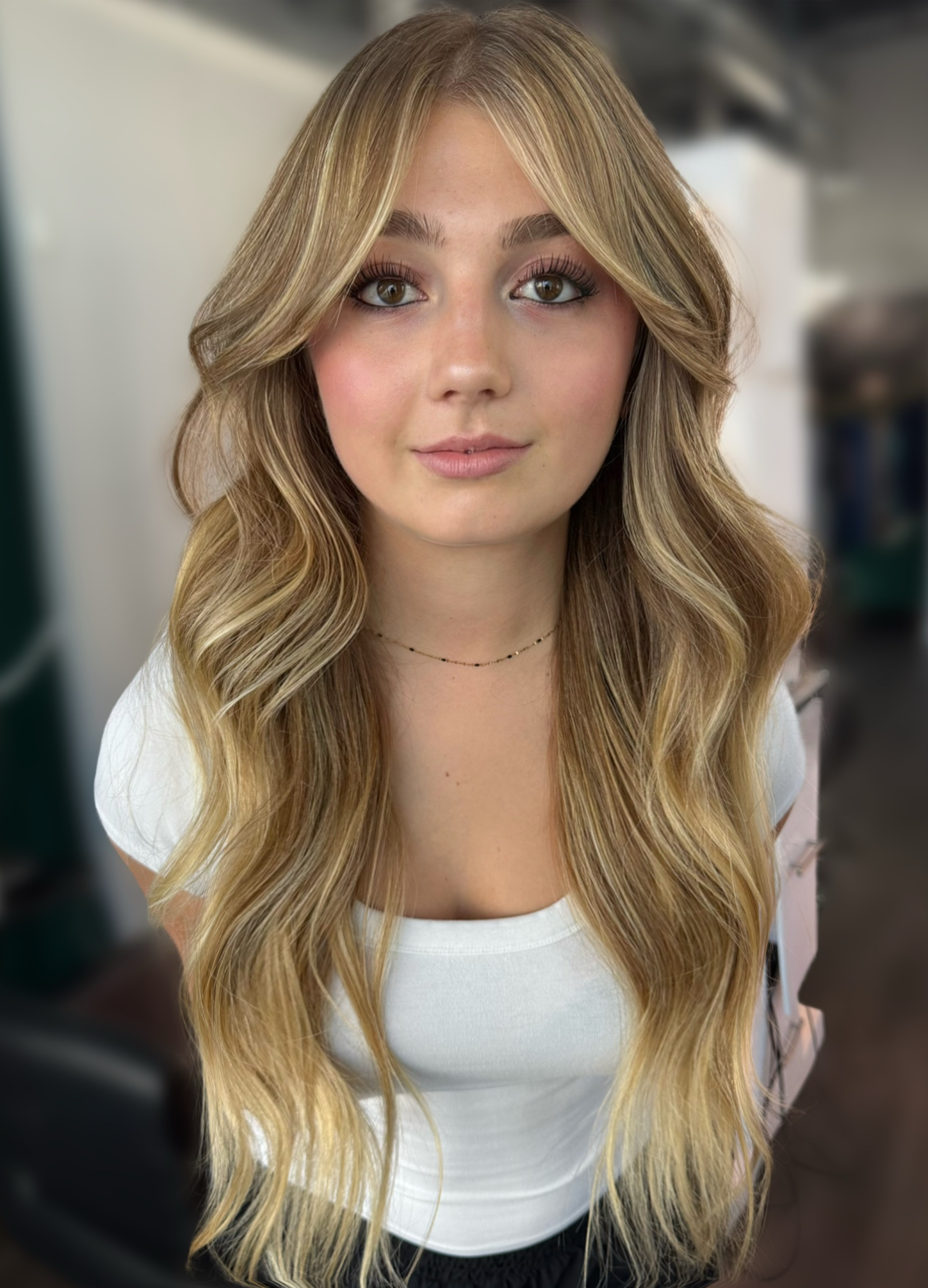 A young woman with long, wavy blond hair and hazel eyes, wearing a white top and a delicate necklace, standing in an indoor setting.