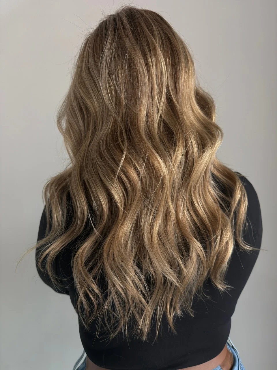 Back view of a woman with long, wavy, light brown hair with highlights, wearing a black top.