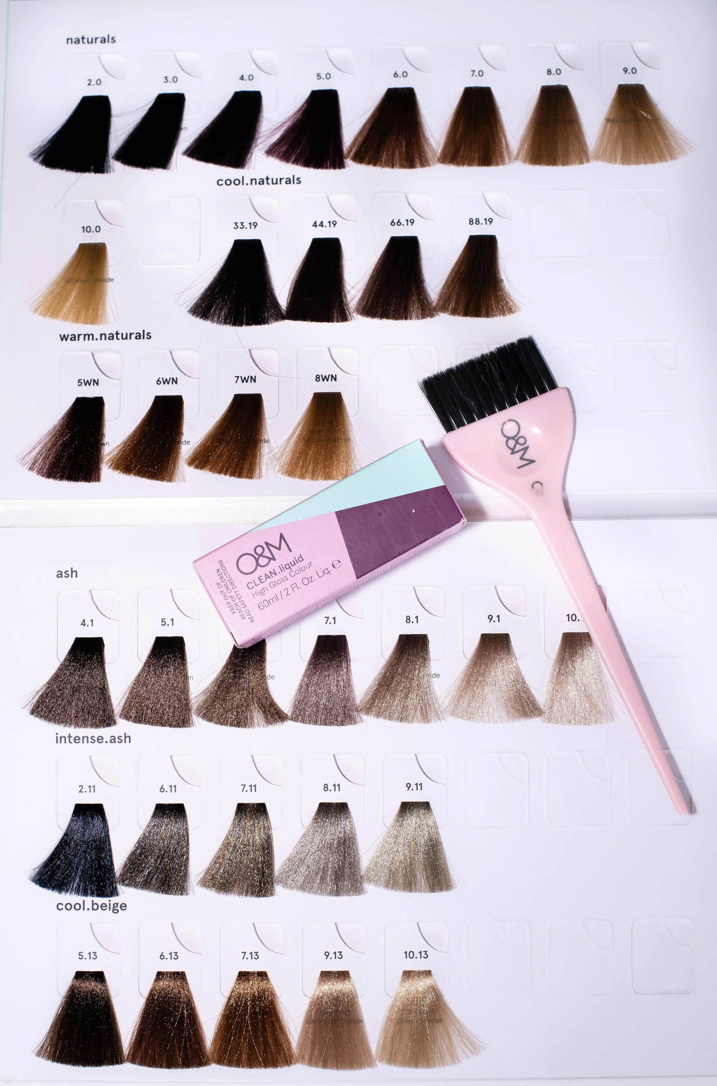 Swatch chart with various hair color samples organized in rows labeled naturals, cool naturals, warm naturals, and intense ash, with hair color names and codes, alongside a hair color brush and a box of O&M hair dye.