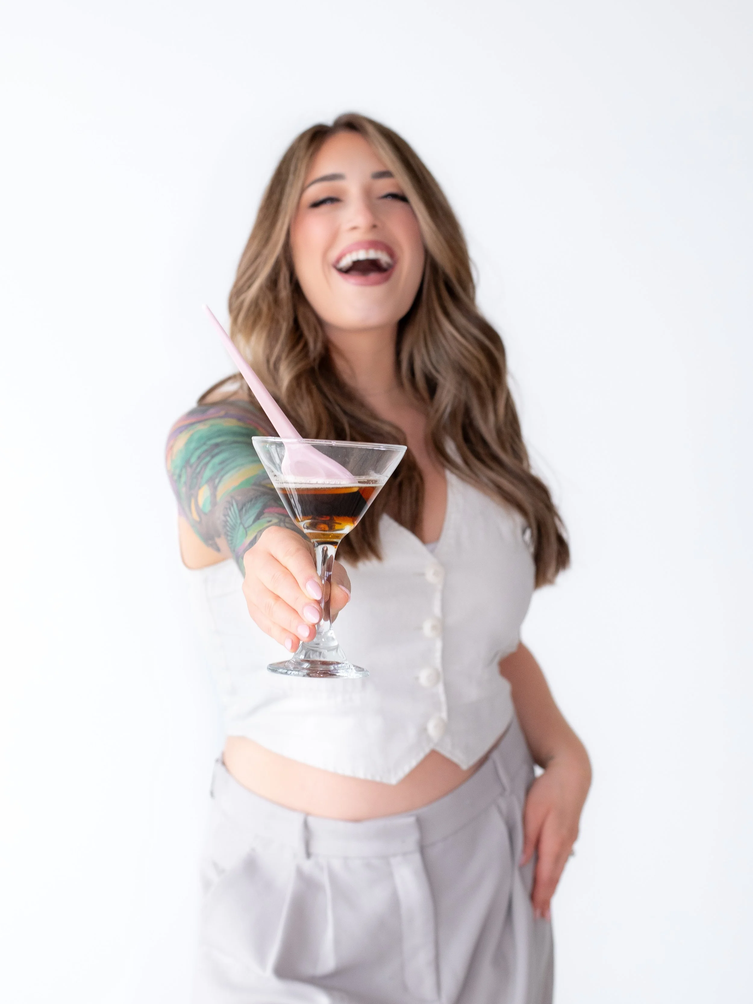 Woman with long brown hair, tattooed arm, in a white buttoned top, holding a martini glass with dark liquor and pink straw, smiling and extending the glass towards the camera, standing against a white background.