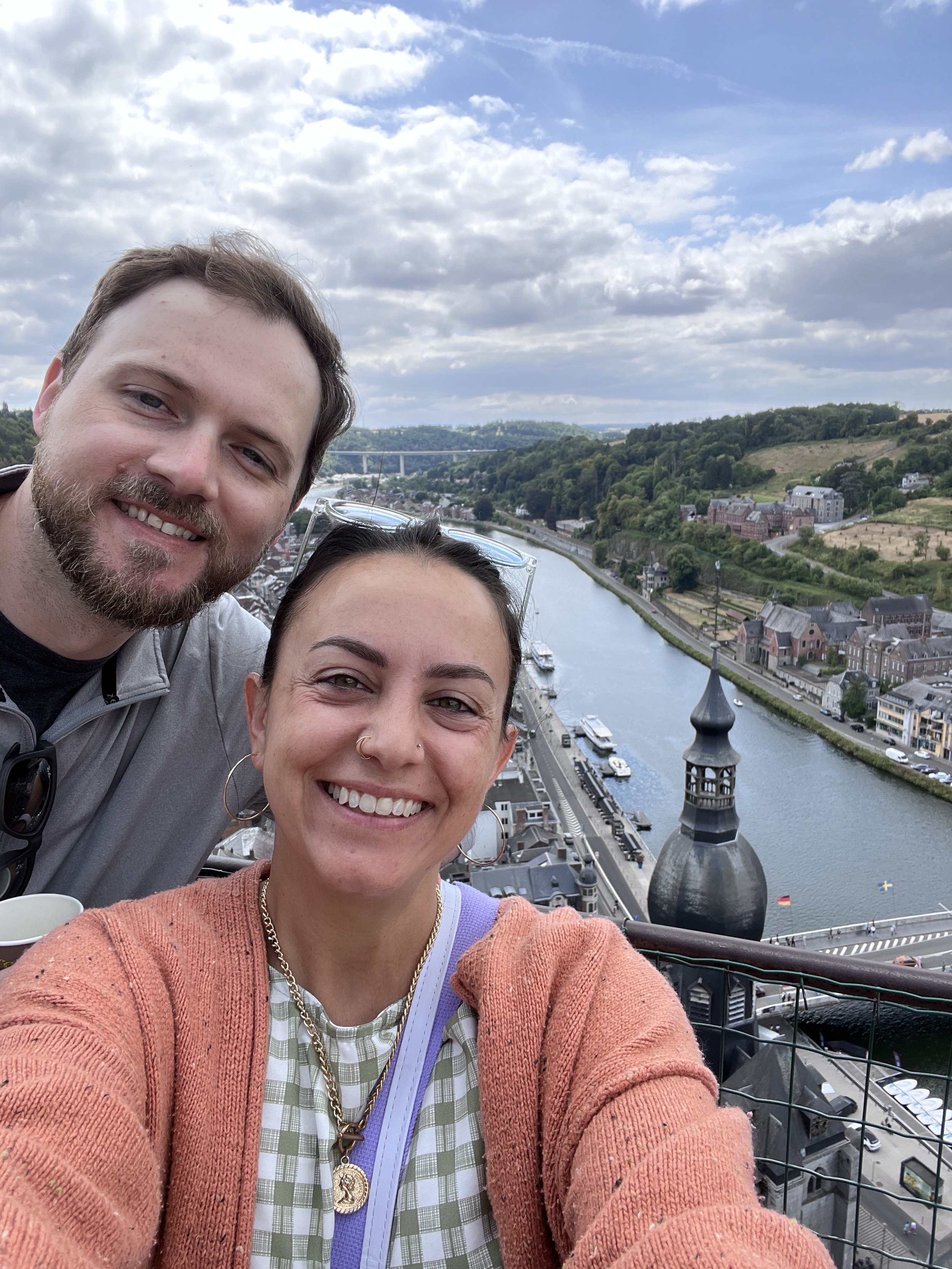 Two smiling people taking a selfie on a high vantage point overlooking a river, with a town and green hills in the background, under a partly cloudy sky.