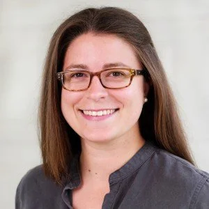 A woman with long brown hair, wearing glasses and a dark gray shirt, smiling at the camera.