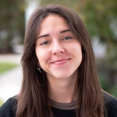 Portrait of a young woman with long brown hair, smiling outdoors.