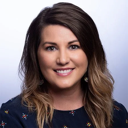 A woman smiling with shoulder-length brown hair, wearing a dark top with small colorful patterns, against a light gray background.