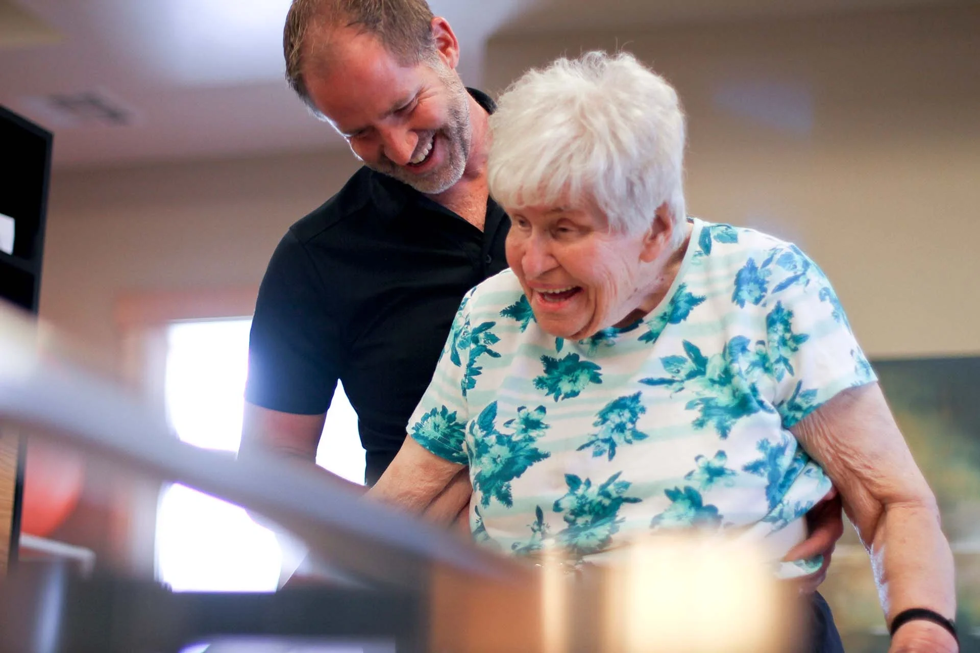 A caregiver assisting an elderly woman with white hair, both smiling, in a bright indoor space.