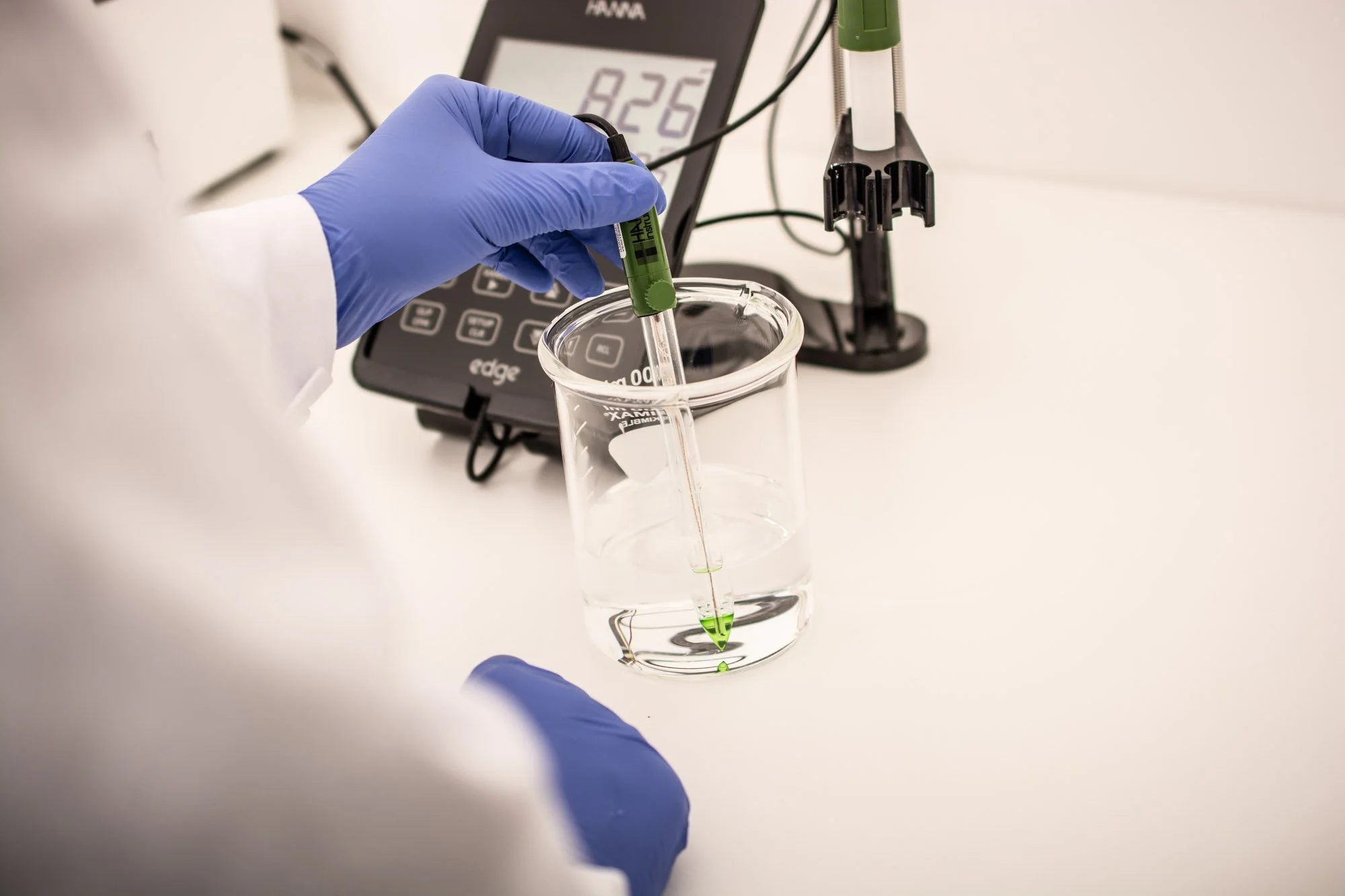 Manos con guantes de látex depositando líquido verde en un vaso medidor, en un laboratorio, con equipo de medición digital en el fondo.