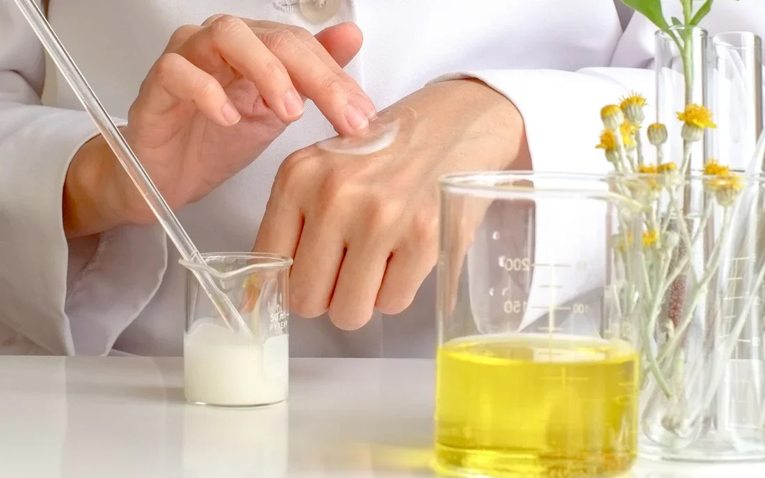 Persona aplicando crema en el dorso de la mano en un entorno de laboratorio o salón de belleza, con frascos y flores en el fondo.
