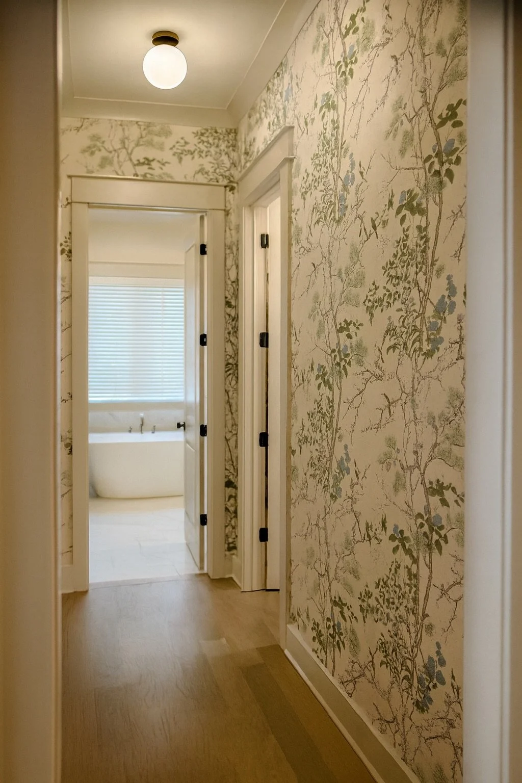 View of a hallway leading to a bathroom with a freestanding bathtub. The walls have floral wallpaper and there is a window with closed blinds in the bathroom. The flooring is wooden.