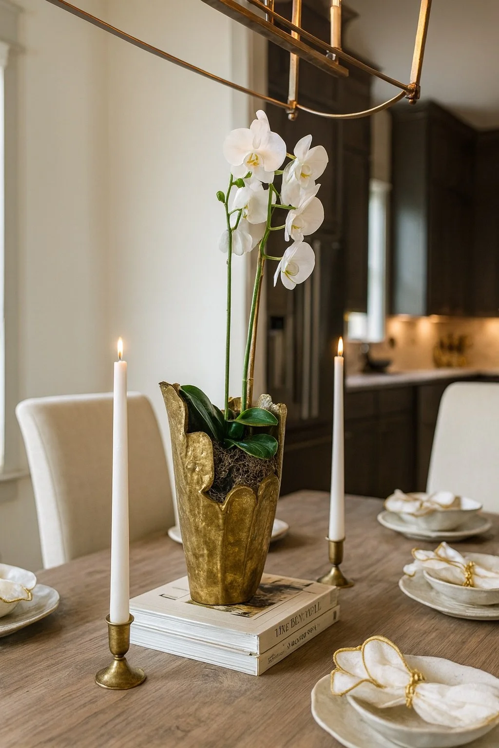 A dining table decorated with a potted white orchid, two tall white candles in brass holders, and white napkins with gold trim placed on plates. The table is in a modern kitchen with dark cabinets.