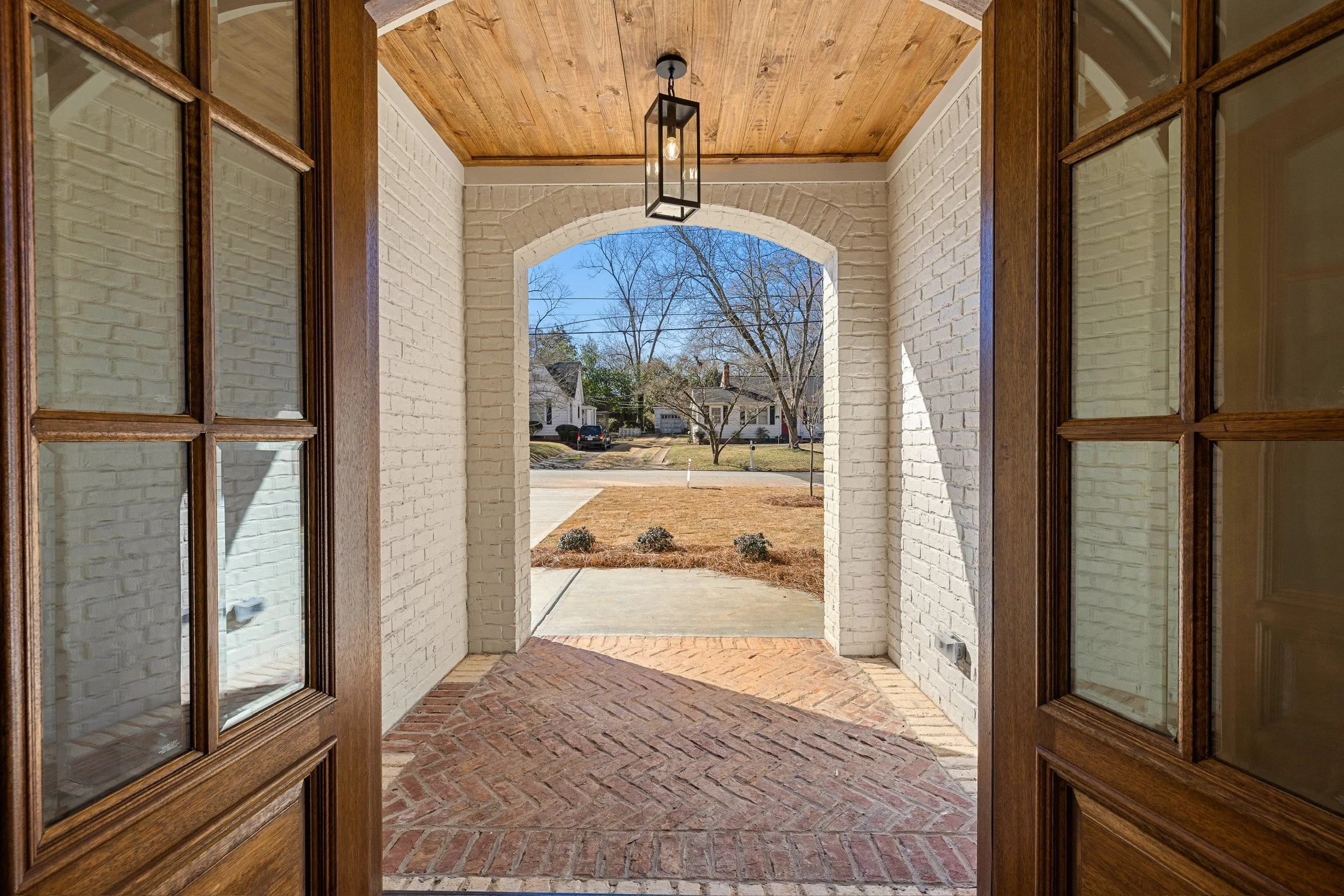 View from a porch with brick flooring, white brick walls, and a wooden ceiling, looking onto a suburban street with leafless trees and houses.