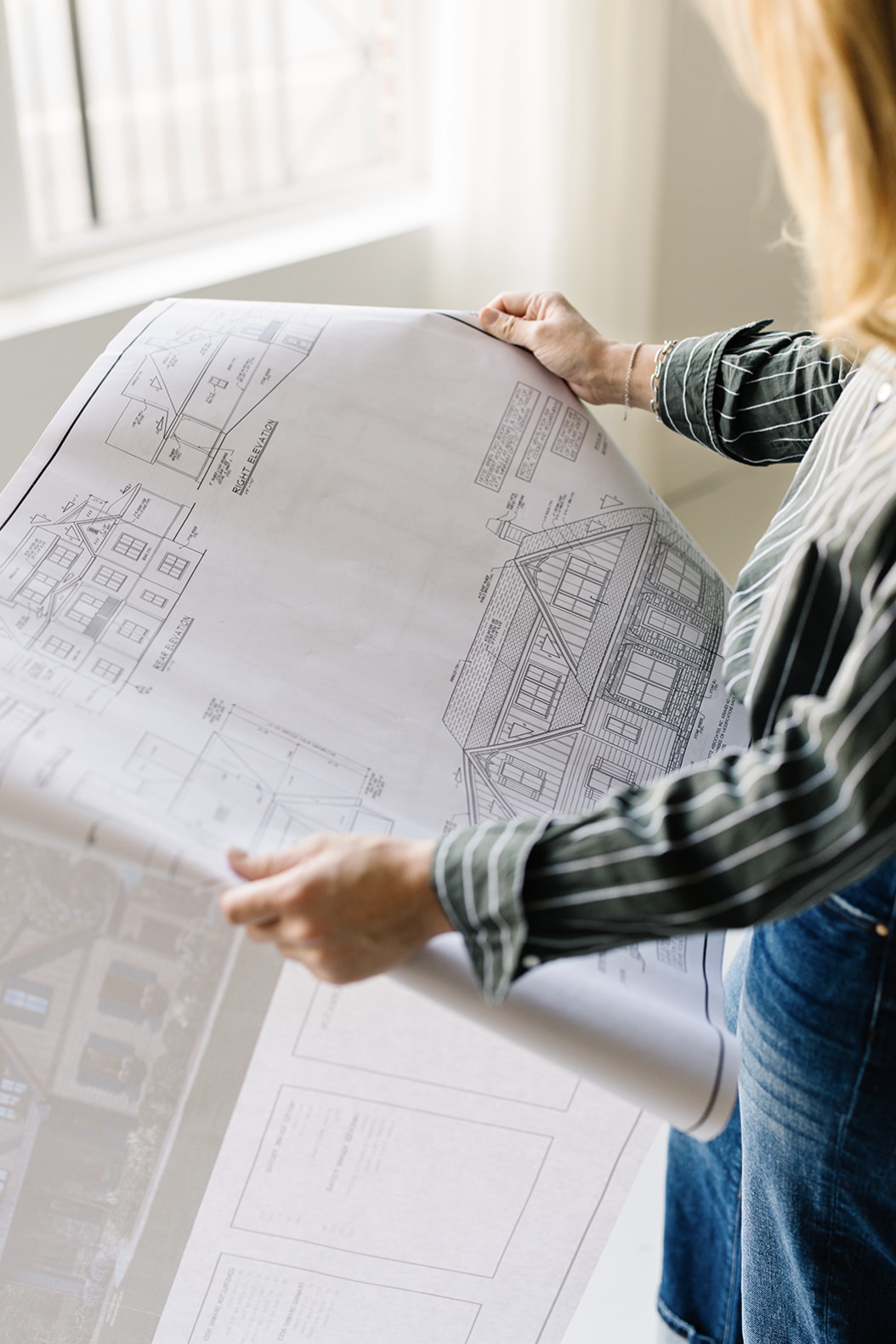 A person holding architectural blueprints of a house, with a window in the background.