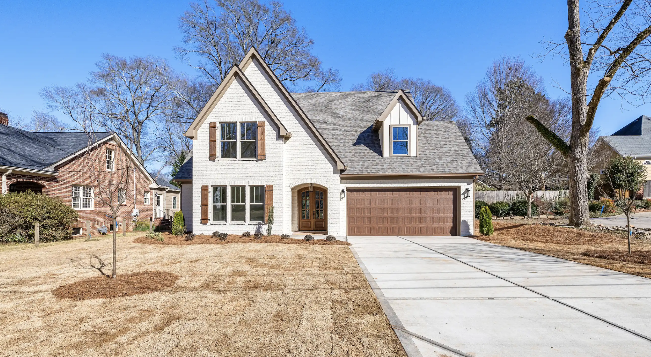 A two-story house with a white brick exterior, brown wooden shutters, and a brown garage door, situated on a lawn with brown grass and a sidewalk leading to the front door, with leafless trees in the background and a bright blue sky.