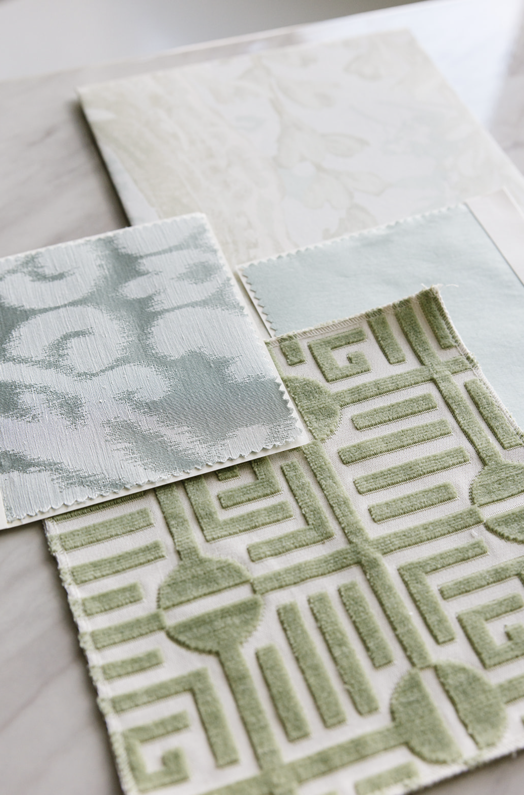 Multiple fabric and wallpaper samples arranged on a marble surface including green geometric patterned rug, pale blue and white fabrics, and light patterned wall coverings.