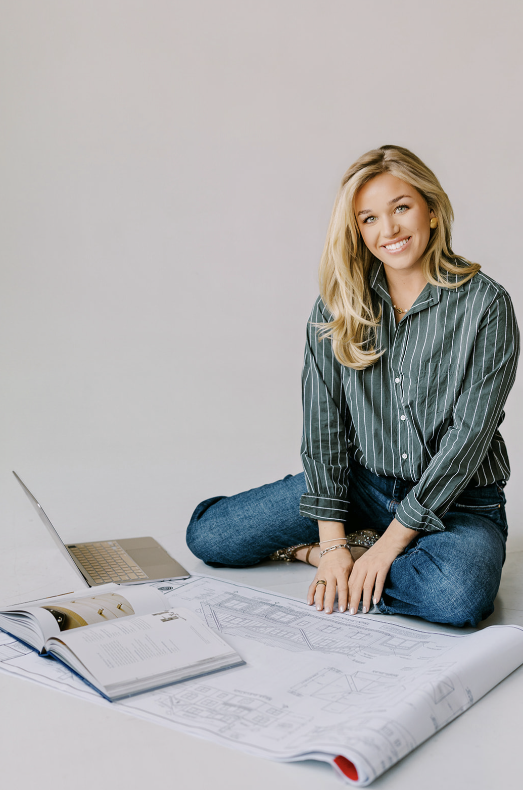 A woman sitting on the floor with rolled-out blueprints, a laptop, and open books, smiling at the camera.