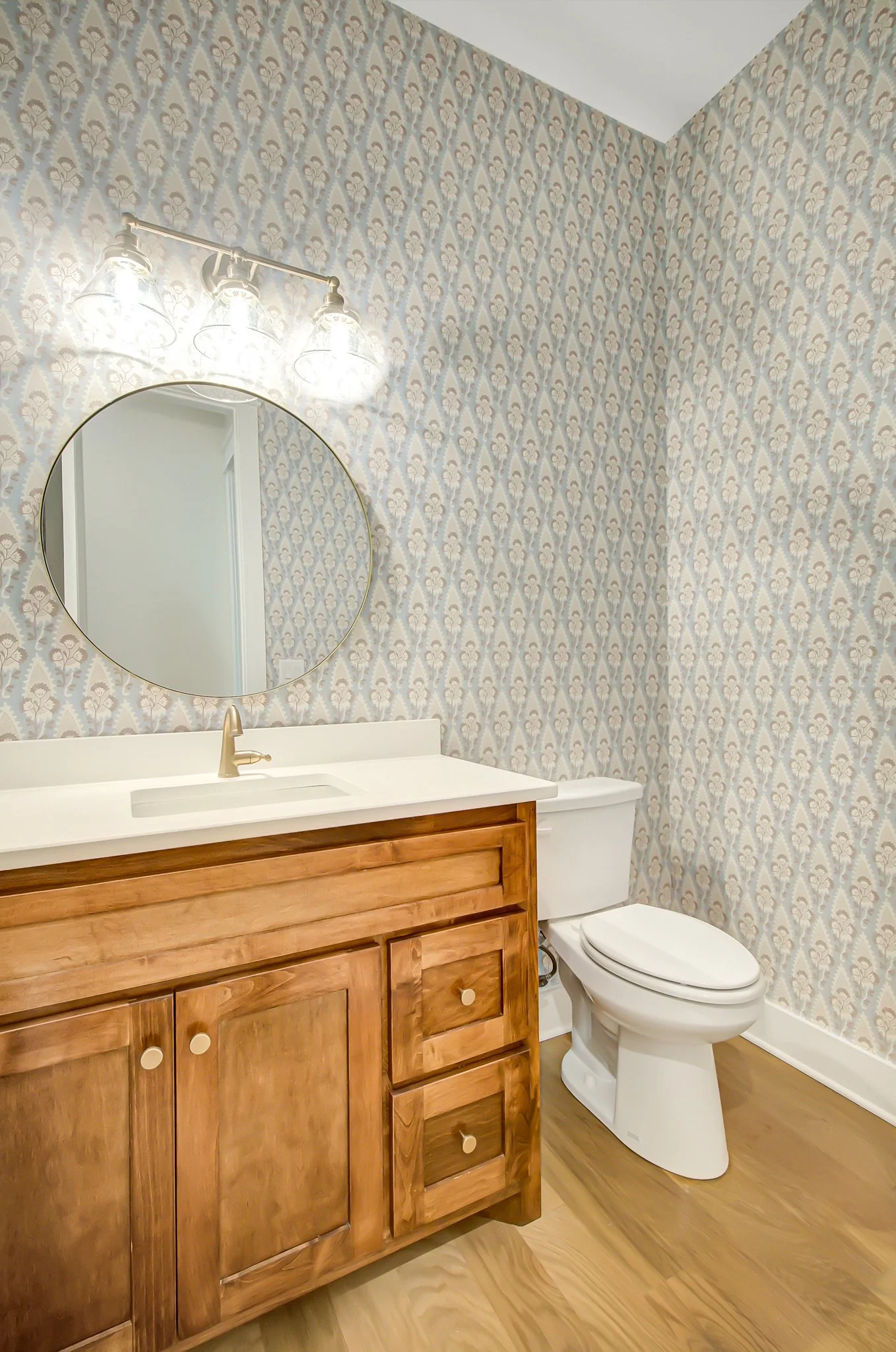 Bathroom with a wooden vanity, a white countertop, an oval mirror, a three-light fixture, and a toilet, with patterned wallpaper and wooden flooring.