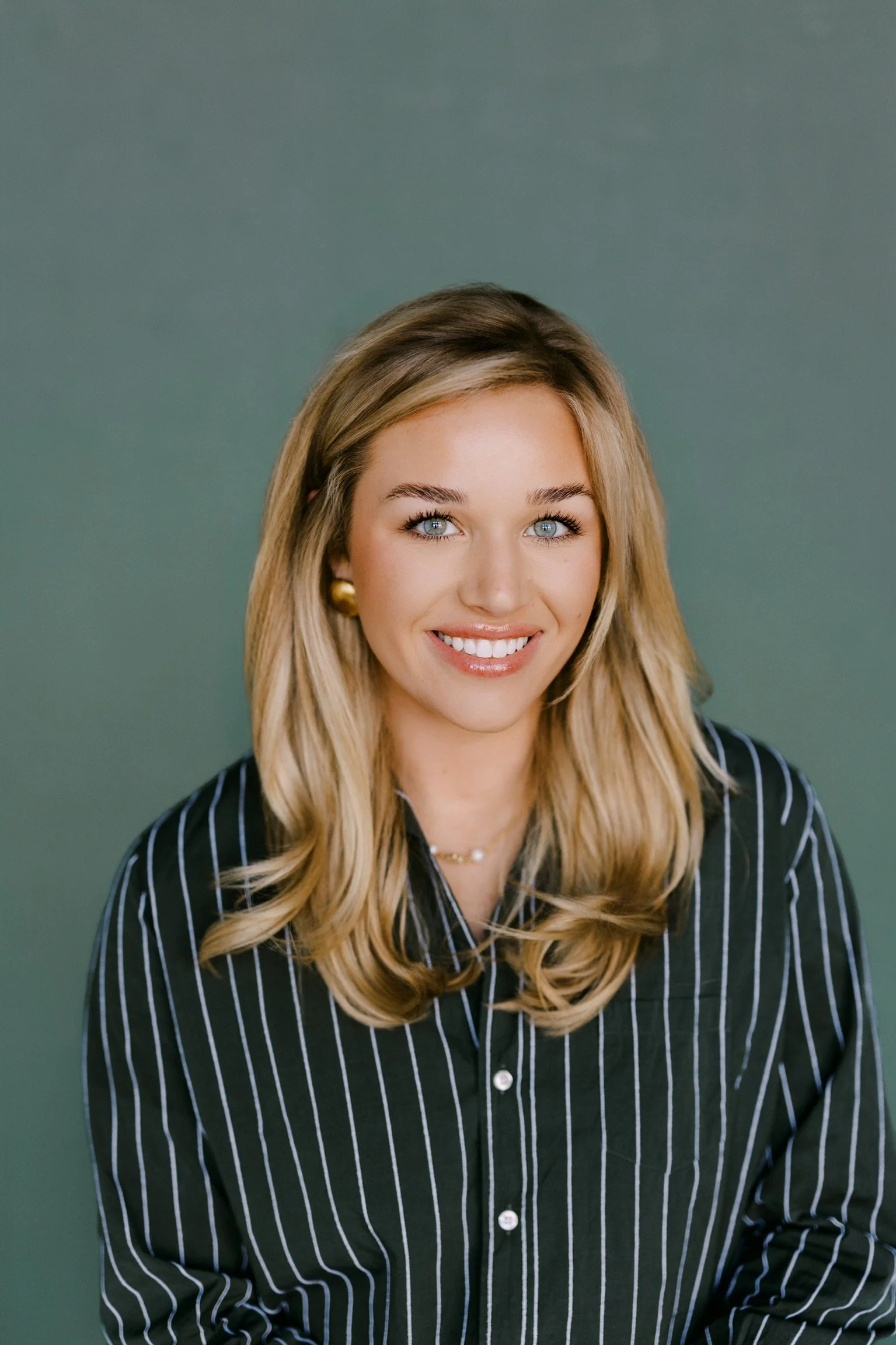 A young woman with blonde hair, blue eyes, and a bright smile, wearing a black shirt with white stripes, gold earrings, and a pearl necklace, against a green background.