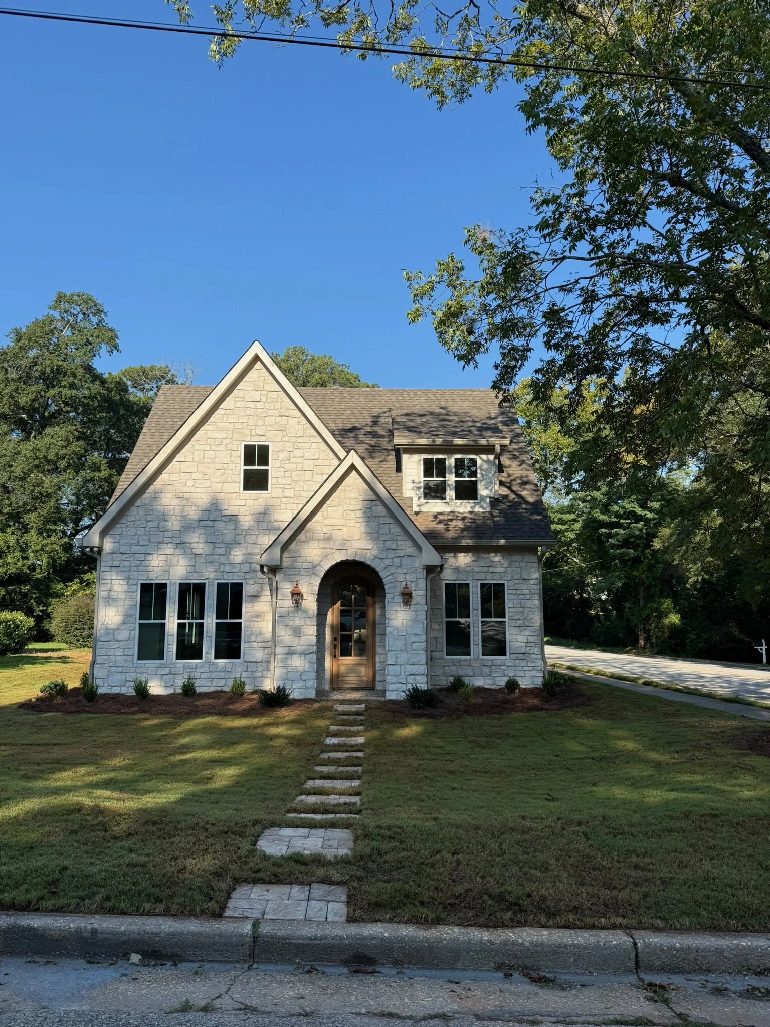 A stone house with a wooden front door and a pathway leading to it, surrounded by a grassy lawn and green trees under a clear blue sky.