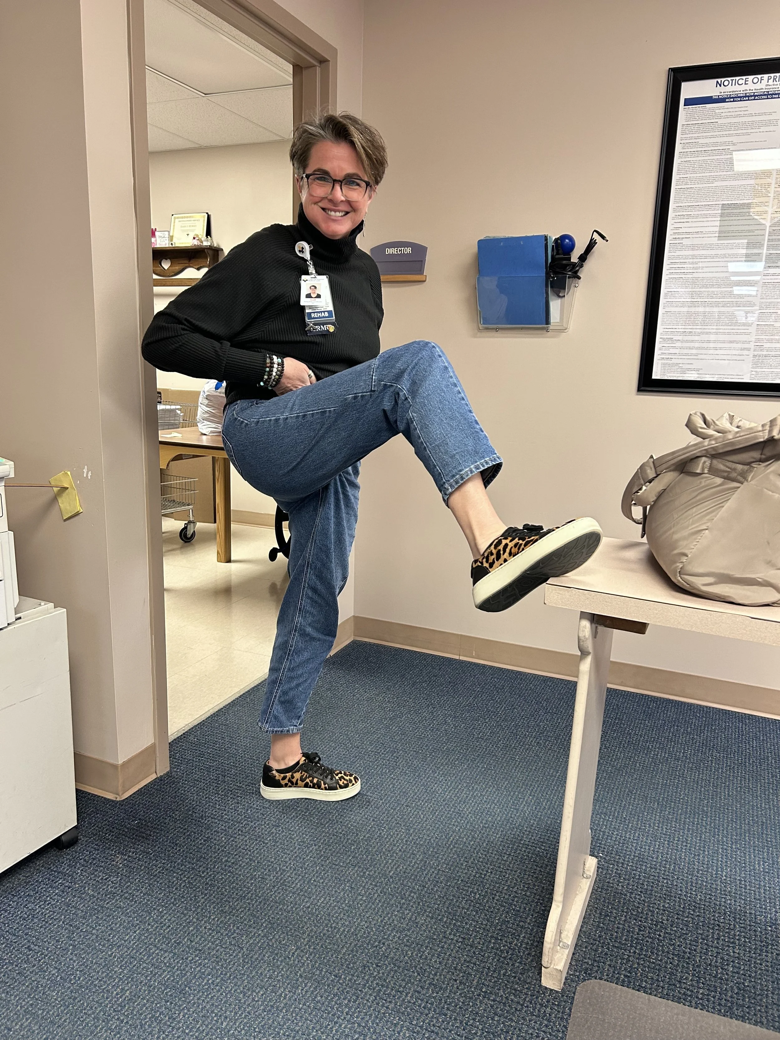 Gail Billman in an office with gray walls, smiling, wearing glasses, a black turtleneck sweater, blue jeans, and leopard print sneakers, holding her leg up on a desk.